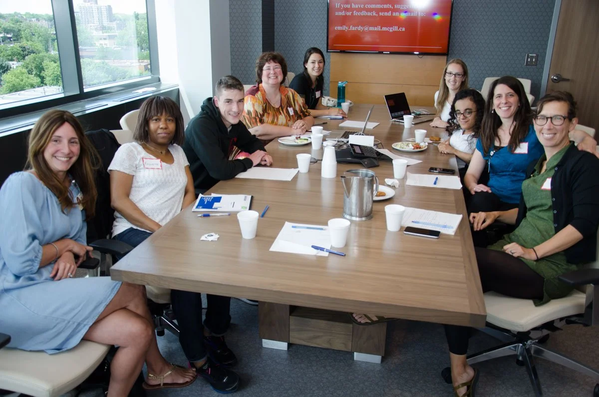 A diverse group of nine women sitting around a conference table in a modern office, smiling for the photo. The table has papers, notebooks, cups, a water pitcher, and plates of food. Large windows reveal a cityscape outside. A large screen on the wal