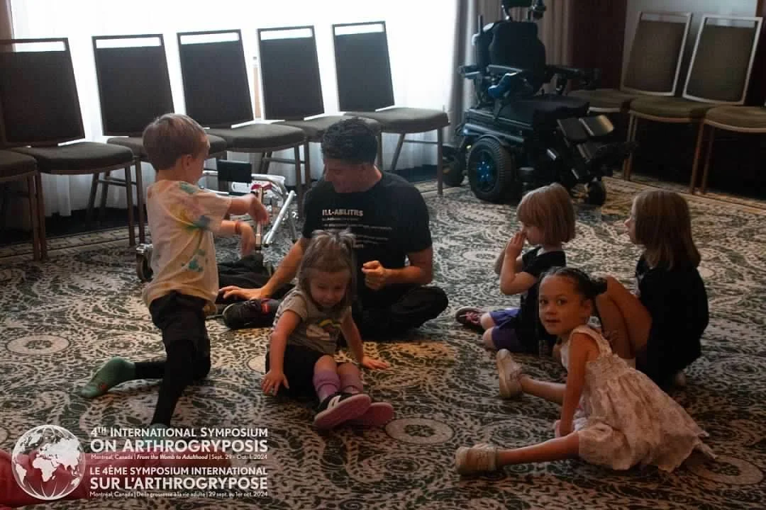 Children and an adult sitting on a patterned carpet in a room with chairs and monitors, with a wheelchair in the background, at the 4th International Symposium on Arthrogryposis.