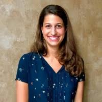 Young woman with long brown hair smiling, wearing a blue patterned blouse, standing against a neutral background.