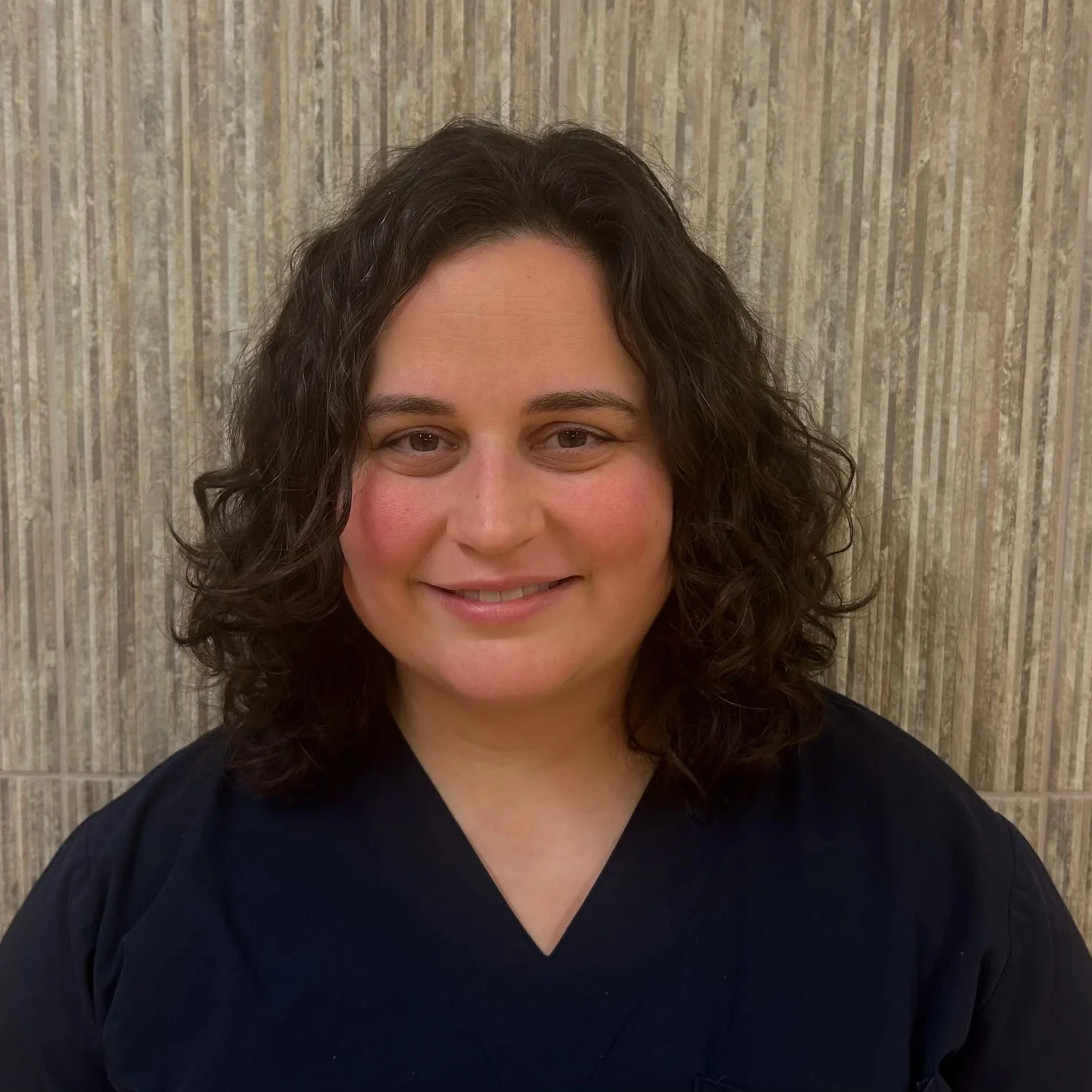 Portrait of a woman with curly dark hair, smiling, wearing a black top, standing against a textured beige background.