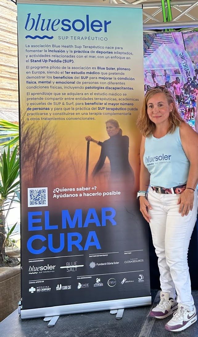 A woman stands next to a large informational banner promoting Blue Soler, a therapeutic stand-up paddleboarding program aimed at improving physical, mental, and emotional health. The banner includes a QR code, logos of supporting organizations, and the phrase "EL MAR CURA" in bold blue letters.