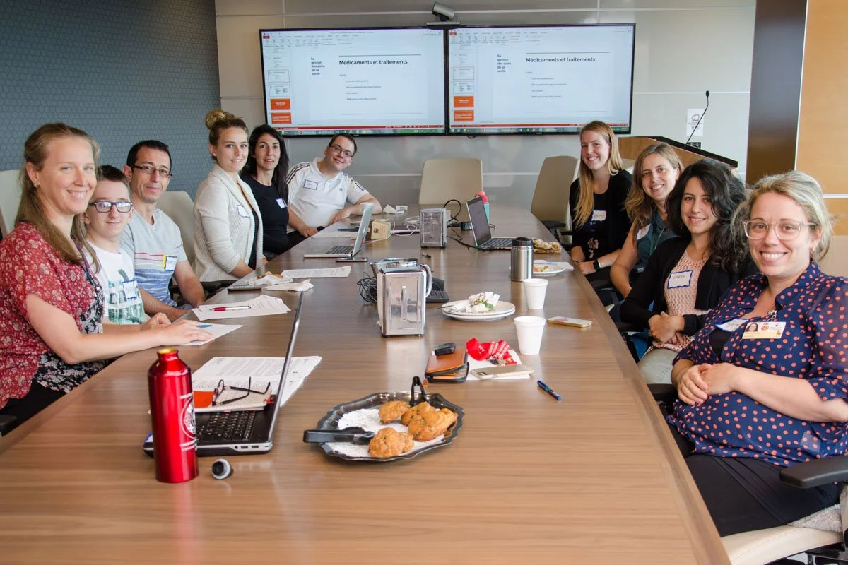 A group of people sitting around a conference table in a meeting room with two screens displaying presentation slides.
