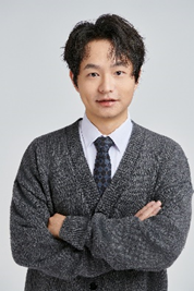 A young man with black, slightly curly hair, wearing a gray sweater over a white collared shirt and a dark tie, standing with his arms crossed against a light gray background.