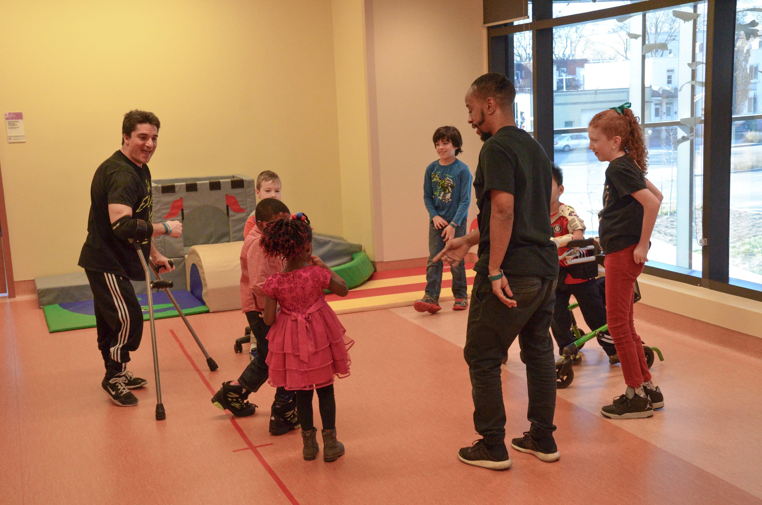 Children playing in an indoor activity room with two adults supervising, with large windows in the background showing an outside view.