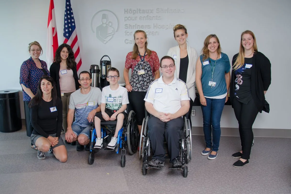 A group of ten people, including children and adults, posing indoors in front of a sign that reads 'Shriners Hospitals for Children'. Some individuals are in wheelchairs, and there are two American flags in the background.