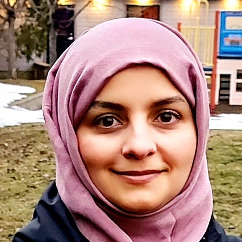 A woman wearing a pink hijab smiling outdoors near a playground and some snow patches.
