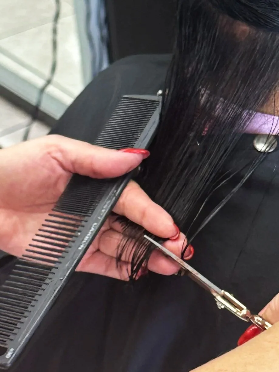 A hairdresser is cutting a person's long, straight black hair with scissors while holding a comb. The person is seated, wearing a black outfit and a purple face mask.