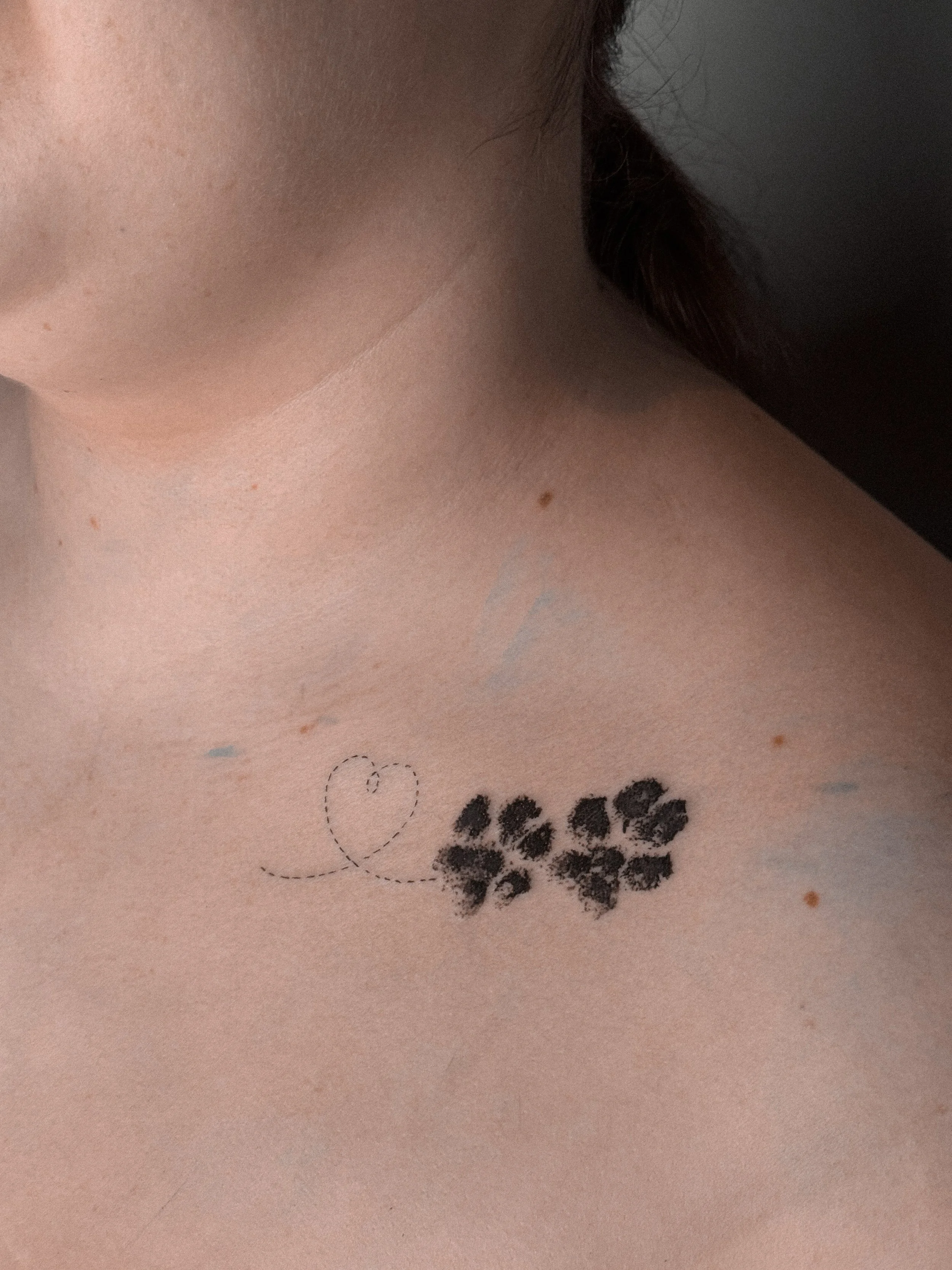 A close-up of a person's shoulder and collarbone with a tattoo of a small heart and a cluster of small flowers.