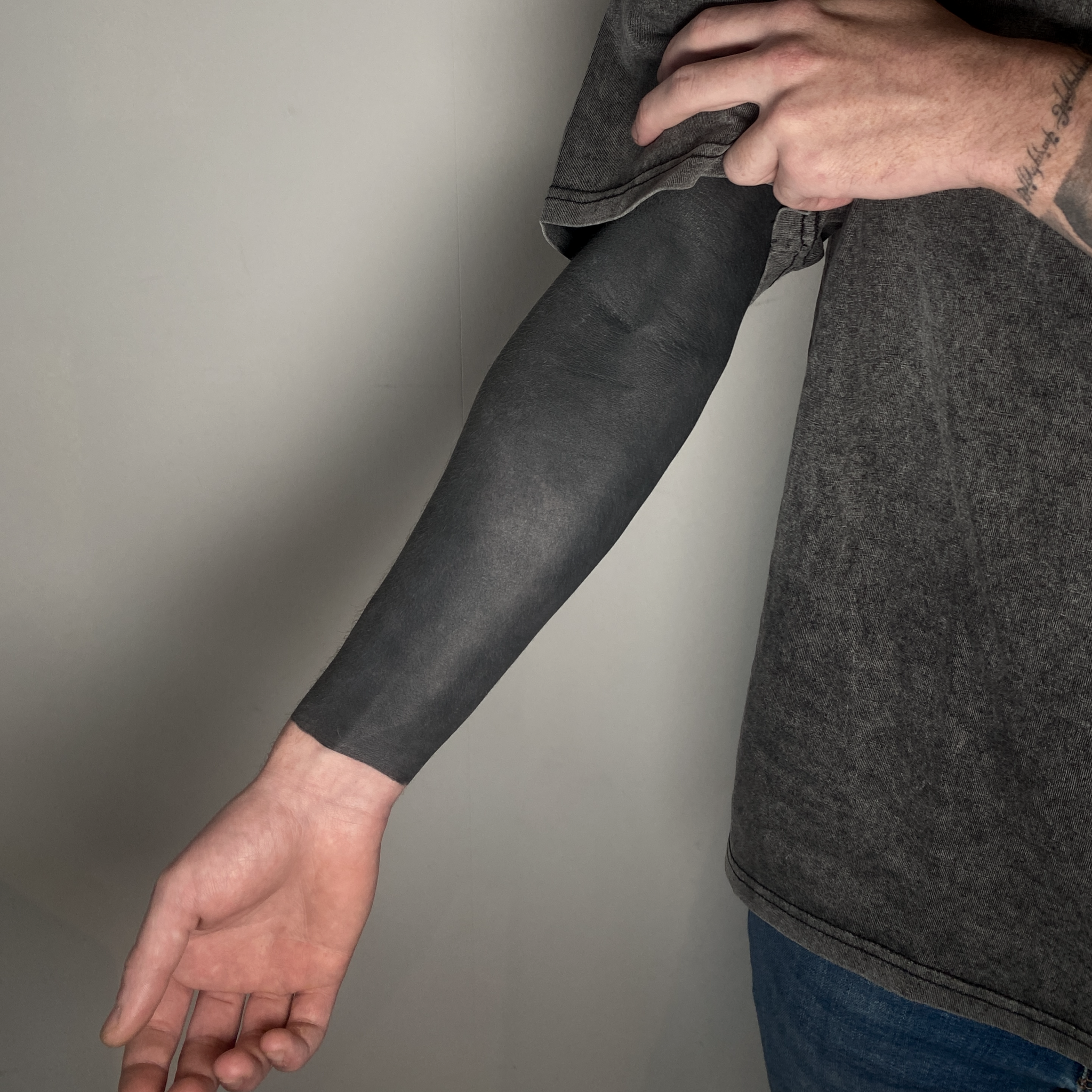 A person is showing their tattooed arm with a black sleeve covering it, wearing a dark gray t-shirt, standing against a plain wall.