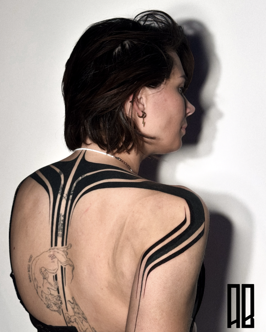 A woman with short brown hair, wearing a black top, with tattoos on her back and shoulder, standing against a white wall. She has a small earring and a necklace, looking to the side with a shadow cast on the wall.