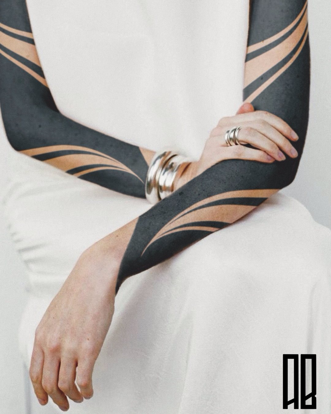 Close-up of a person's arm with black and nude tribal-style tattoos, wearing multiple silver bangles, resting on their crossed arm, against a plain white background.