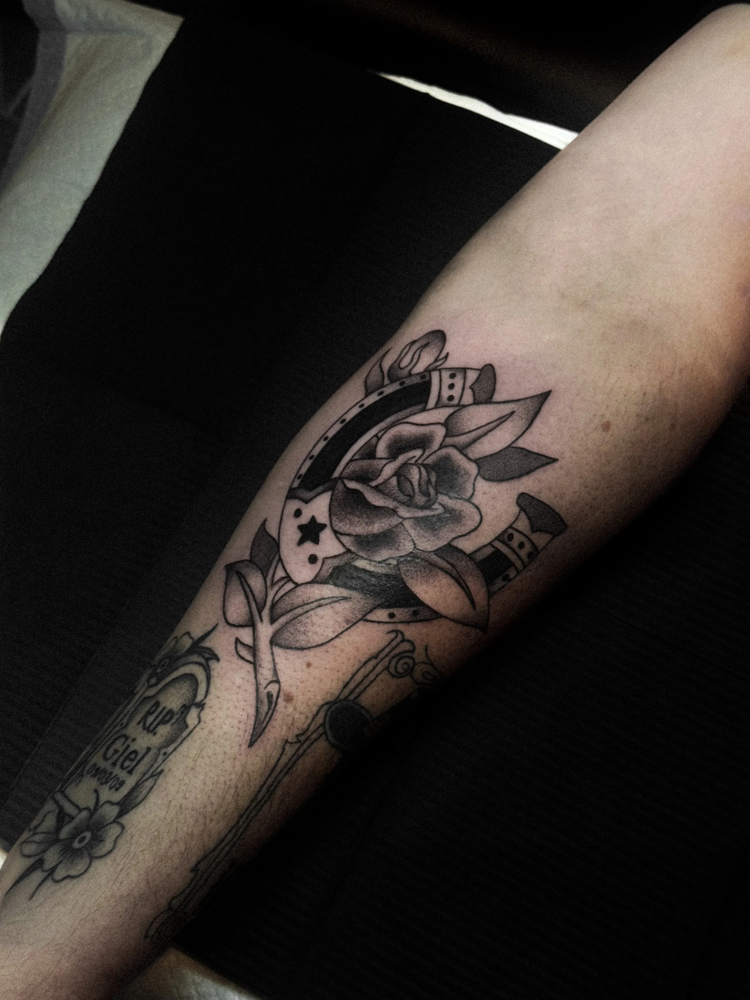 Tattoo of a rose flower with leaves, a stopwatch, a small star, and a memorial plaque with the inscription 'RIP Giel 2009', all in black ink.