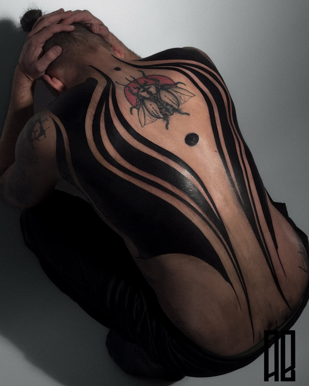 A person with a large back tattoo featuring a bee, black abstract lines, a red semi-circle, and a black dot, seen from behind with their head resting on their knees.