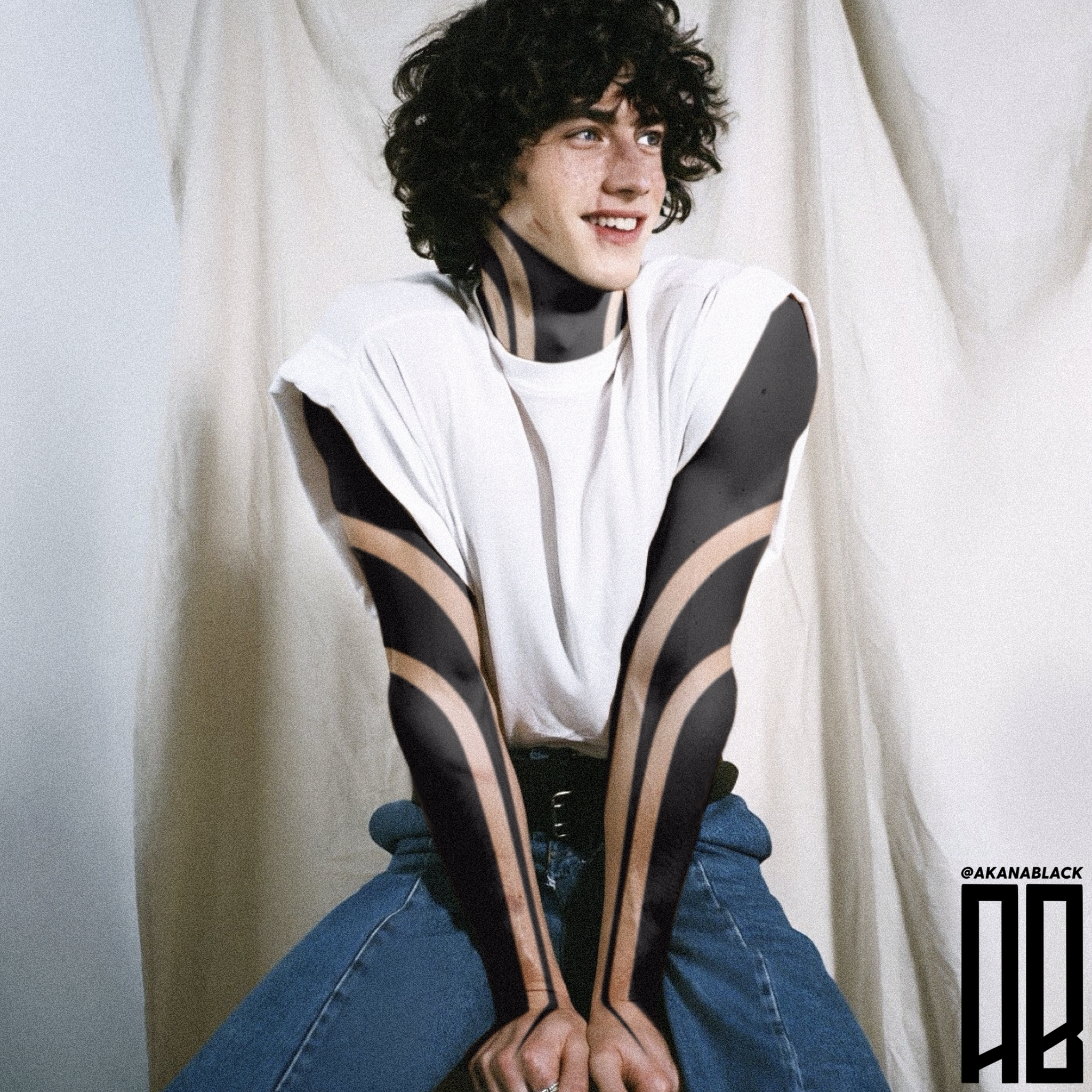 A young man with curly black hair and tattoos on his neck and arms, wearing a white sleeveless shirt and jeans, smiling and sitting with hands clasped forward against a cream-colored curtain background.