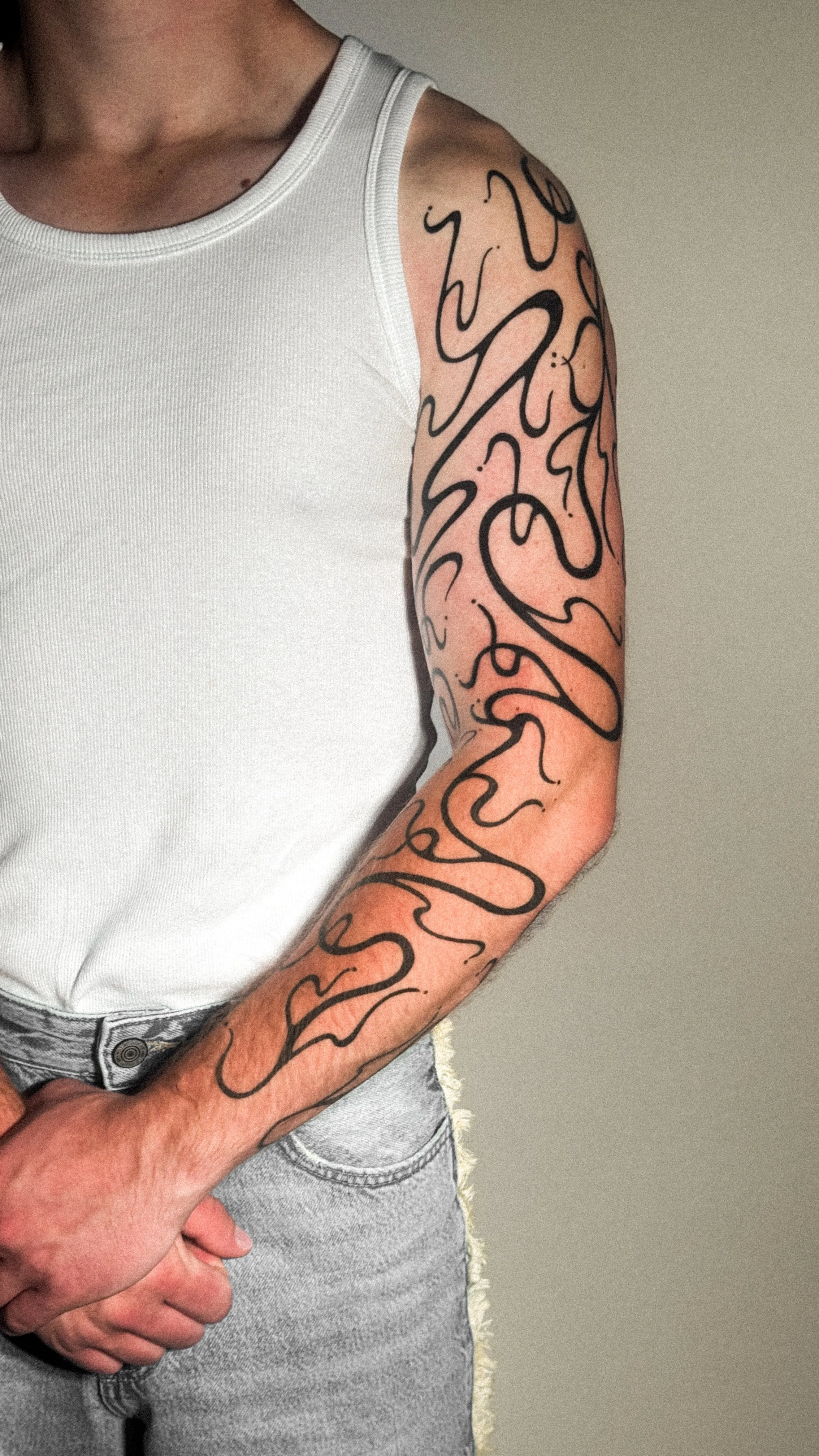 A person wearing a white tank top and light gray jeans with a large tattoo of black swirling lines on the left arm.