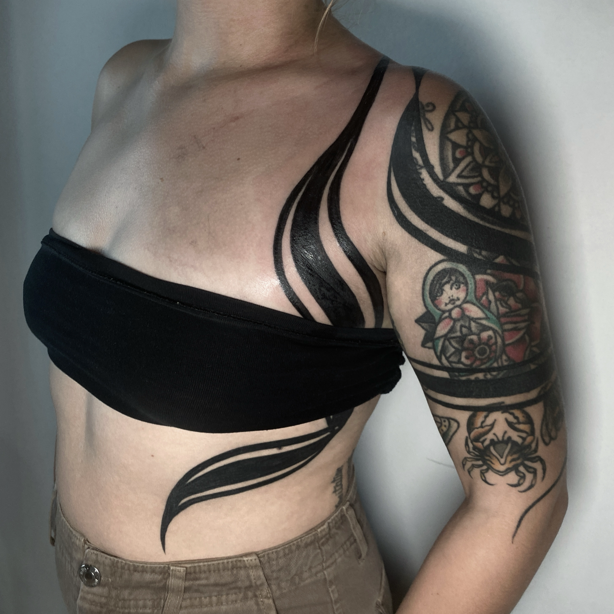 Close-up of a woman's upper body showing tattoos on her shoulder, arm, and side, wearing a black bandeau top and beige pants.