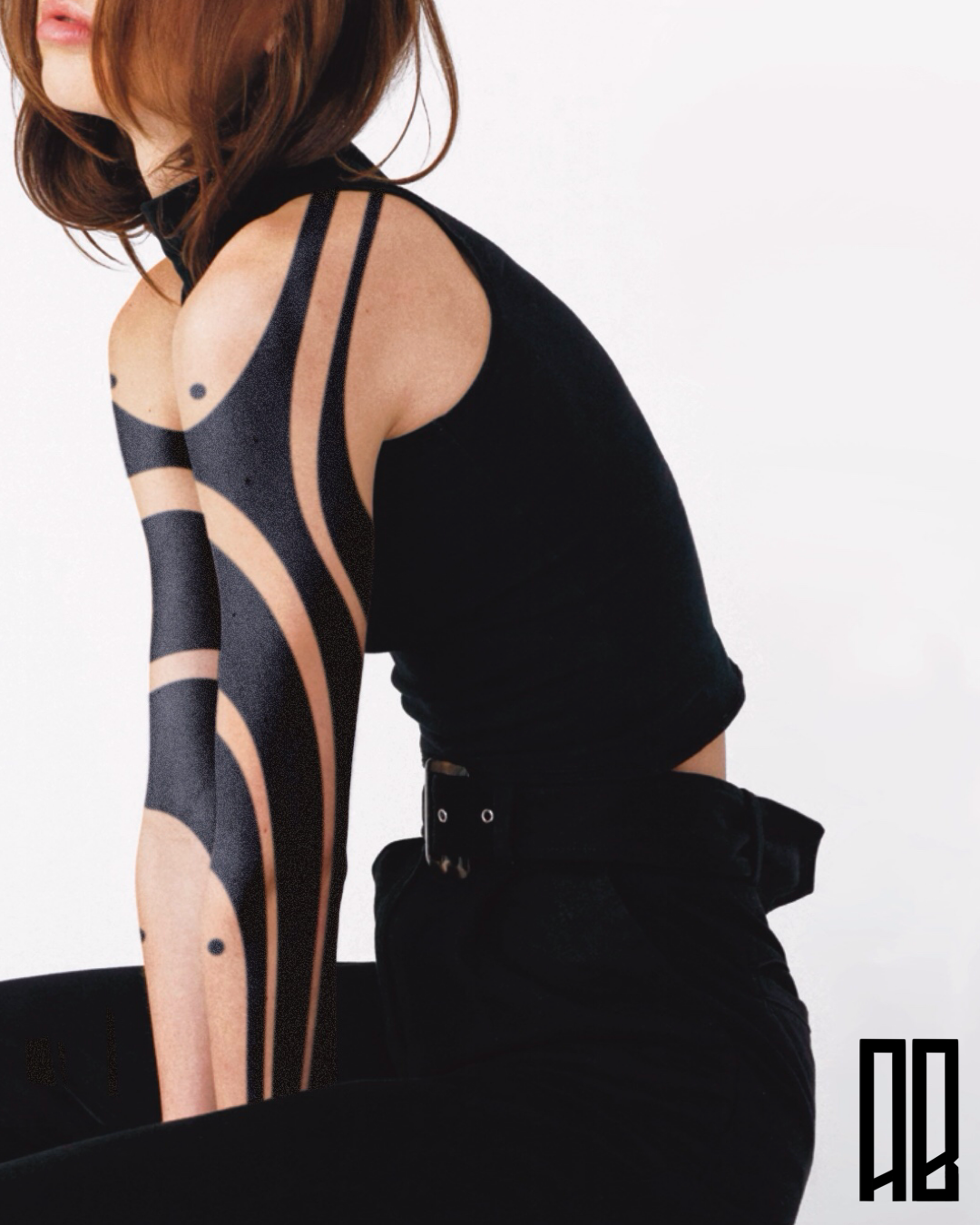 A woman with shoulder-length brown hair wearing a black sleeveless top with cutouts and tattoos on her arm, posing against a plain white background.