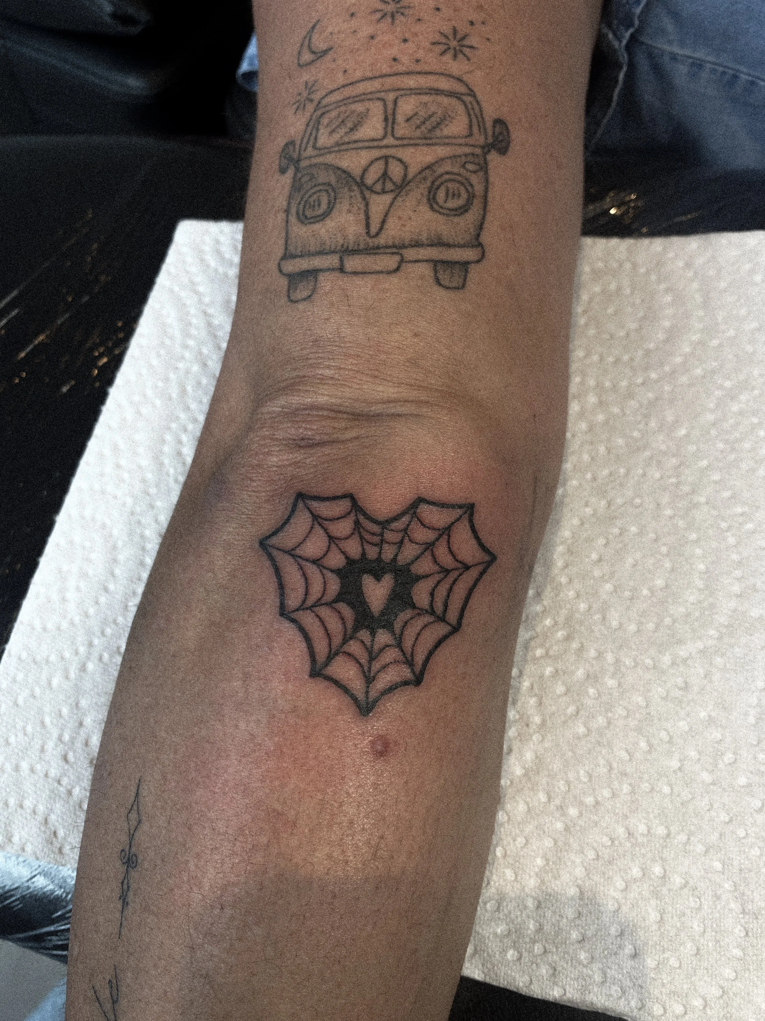 Tattoo of a spider web with a heart in the center on the forearm and a tattoo of a vintage van with a peace sign on the front on the upper arm.