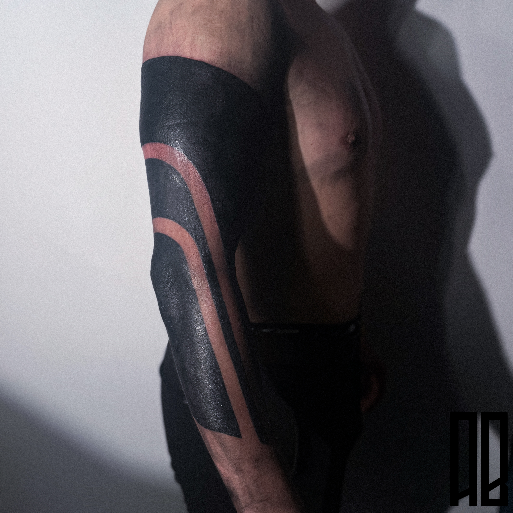 Close-up of a person's upper body with a covered eye, showing a black and brown arm tattoo, and a bare chest against a plain background.