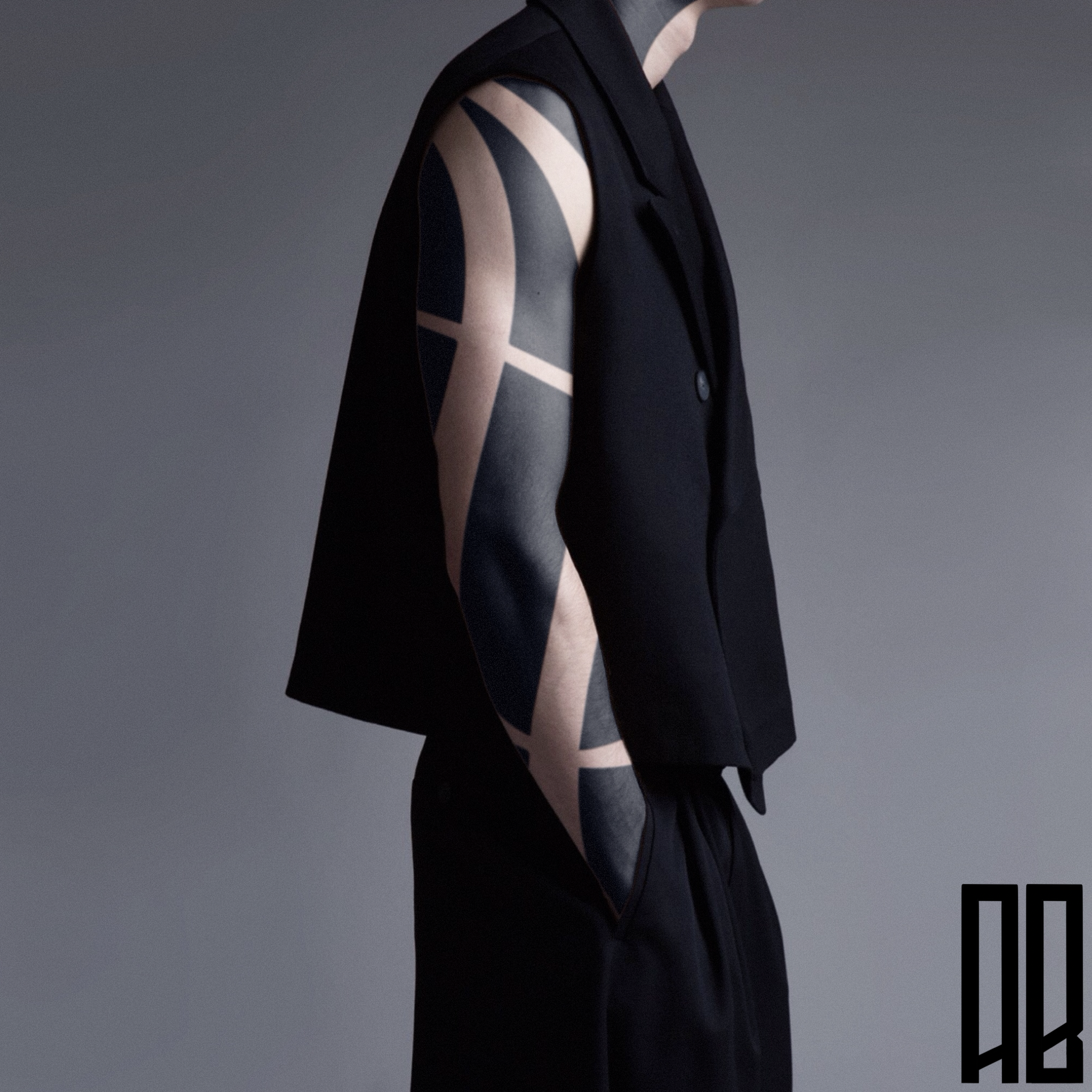 Person wearing a sleeveless black vest and black pants, with a geometric black and beige tattoo on their arm, standing against a plain gray background.