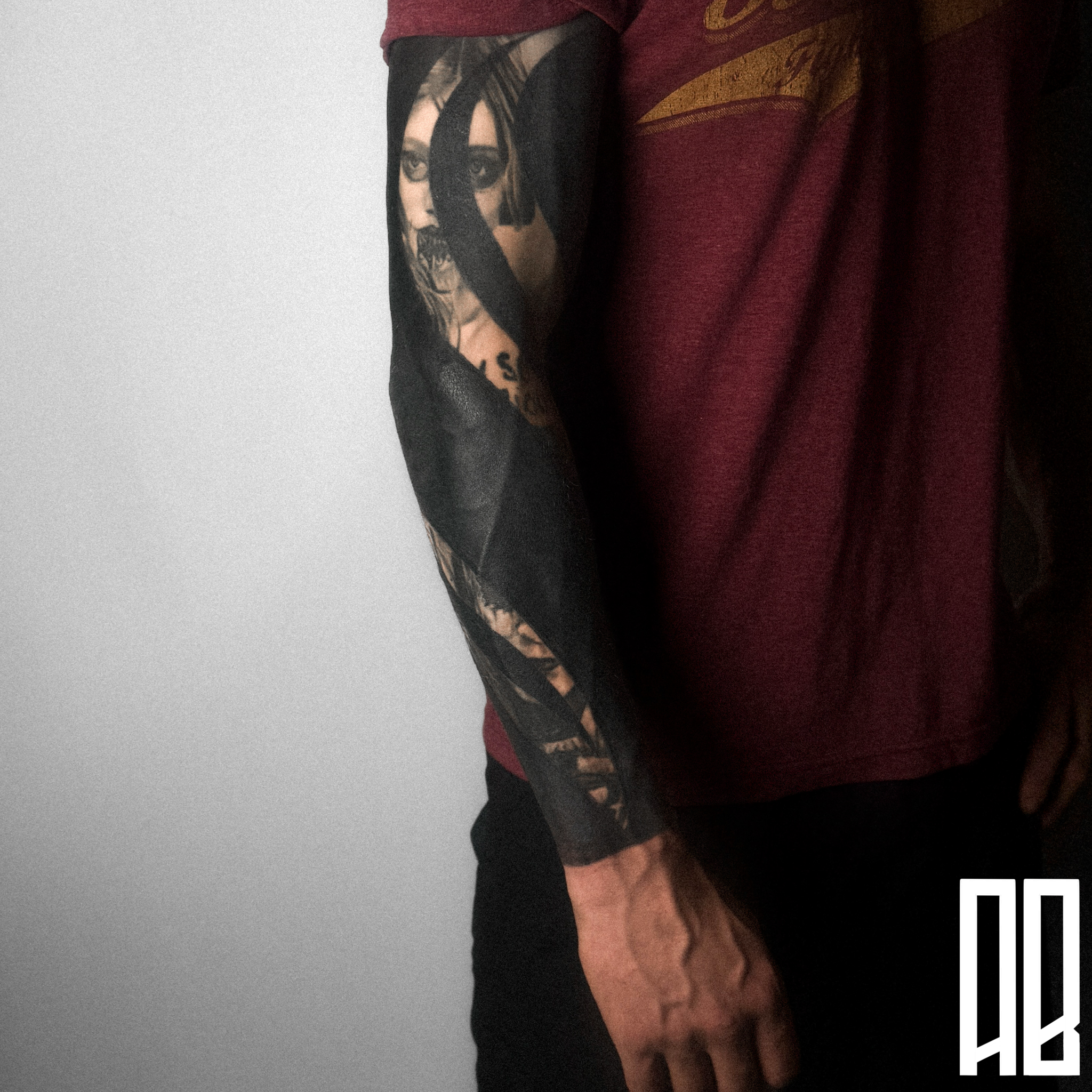 A person's arm with a detailed black and gray tattoo of a woman's face wearing glasses, with long hair, on the forearm. The person is wearing a maroon t-shirt and black pants, standing against a plain light gray background.