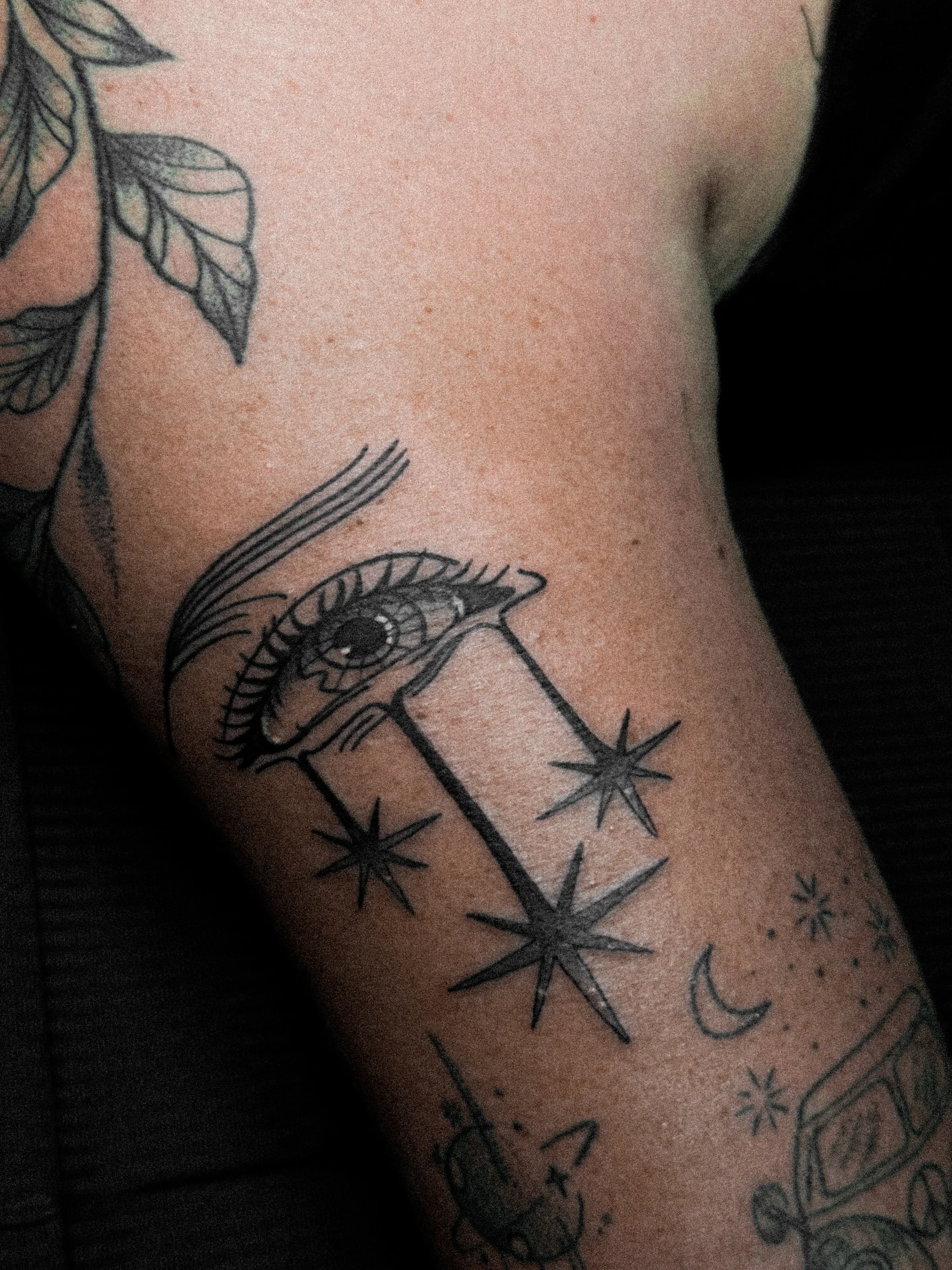 Tattoo of an eye with eyelashes, a spaceship shooting a beam of light, stars, a crescent moon, and a planet on skin.