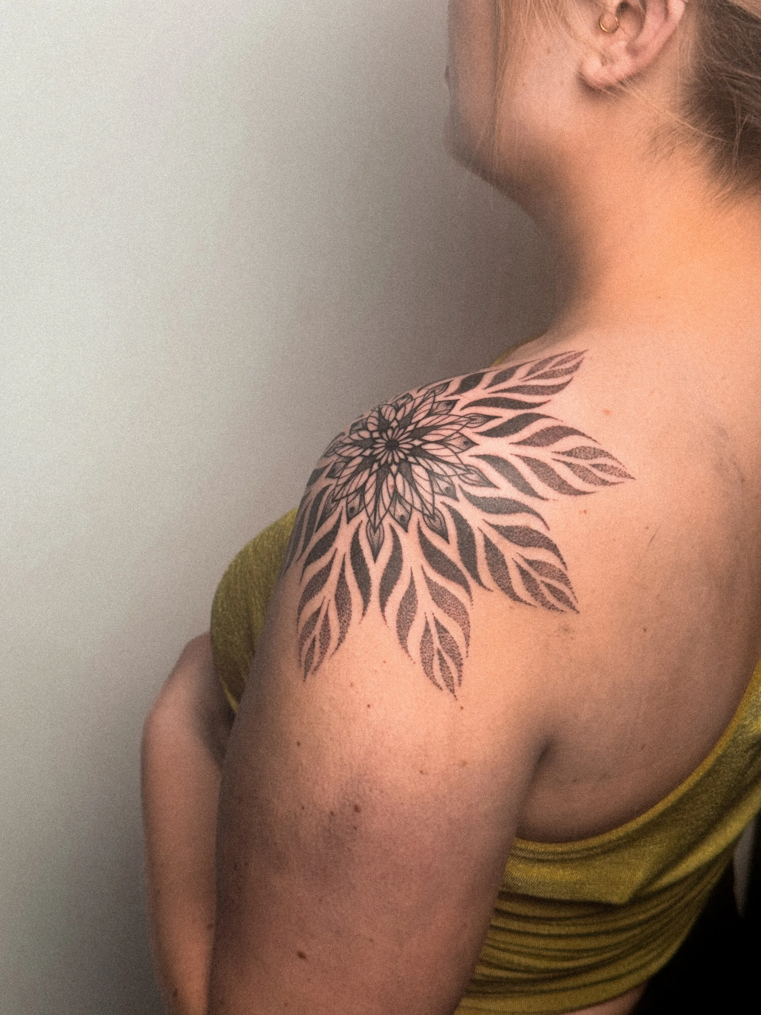 A woman with a large black and grey floral tattoo on her shoulder and upper arm, wearing a yellow sleeveless top.