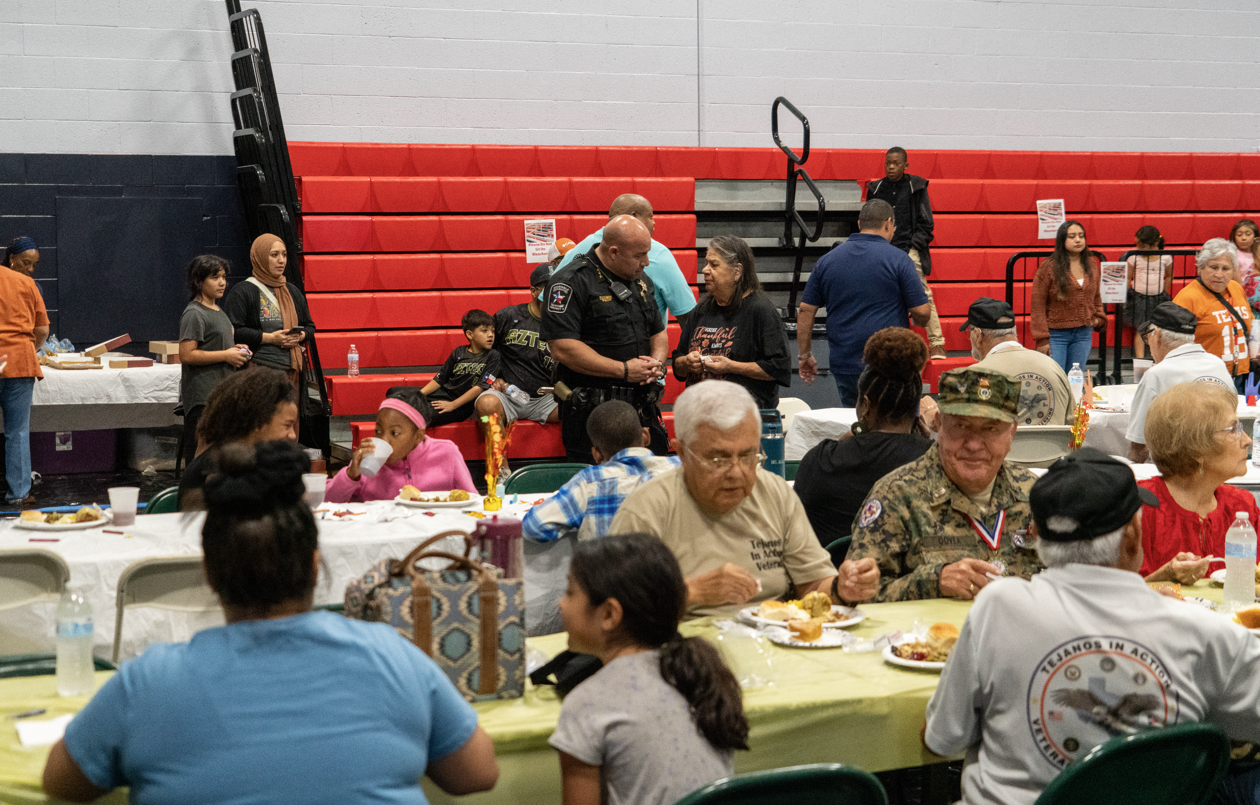 Constable Gabe Padilla at a community gathering at a banquet with people of various ages and backgrounds seated at tables, some eating and others socializing, with a few vendors serving food in the background and others standing near red bleachers.