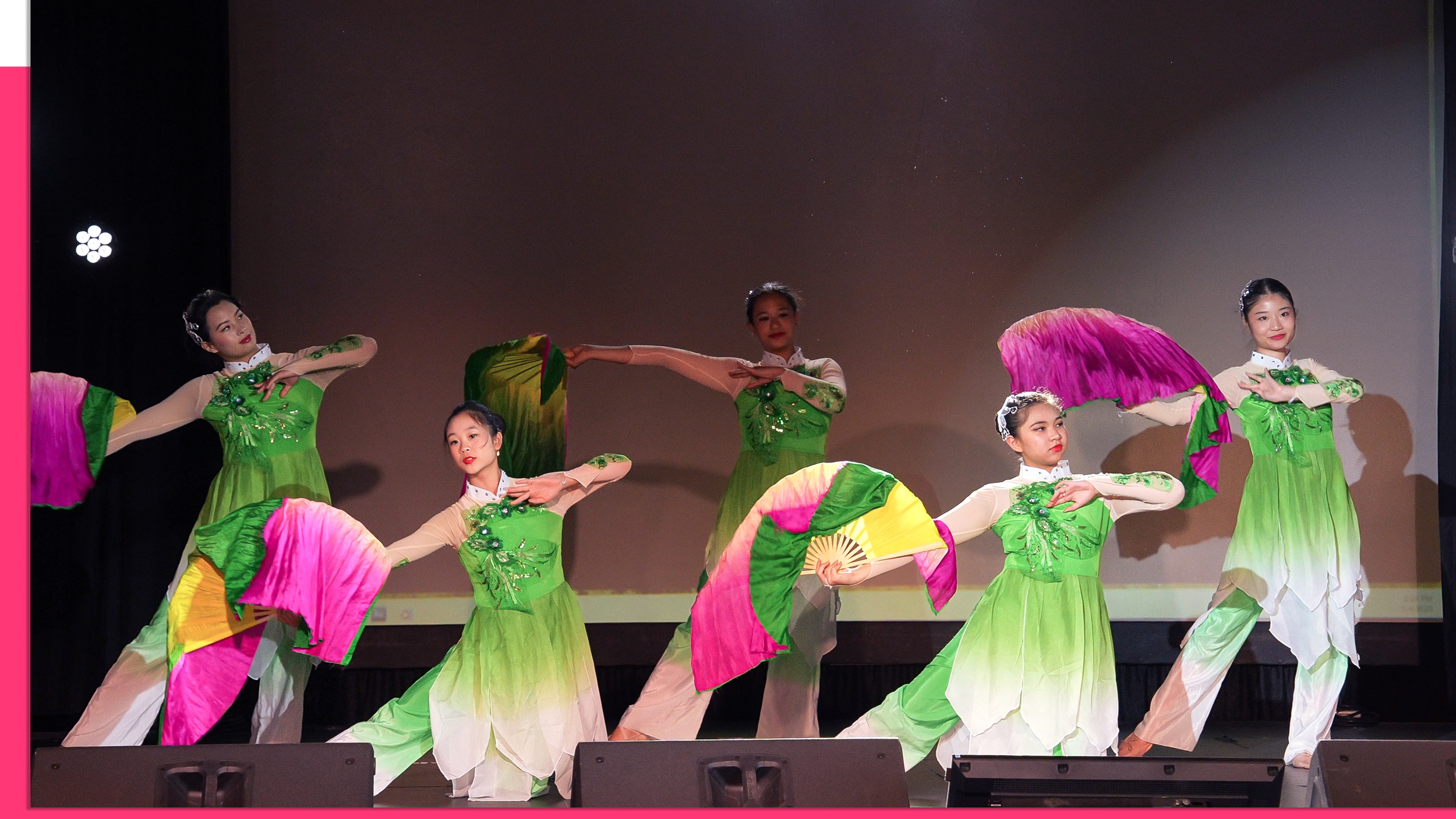 Six young women perform a traditional dance on stage, wearing green and white dresses with floral embroidery, and holding colorful pink and yellow fans. The background screen is plain, and stage lights illuminate the dancers.