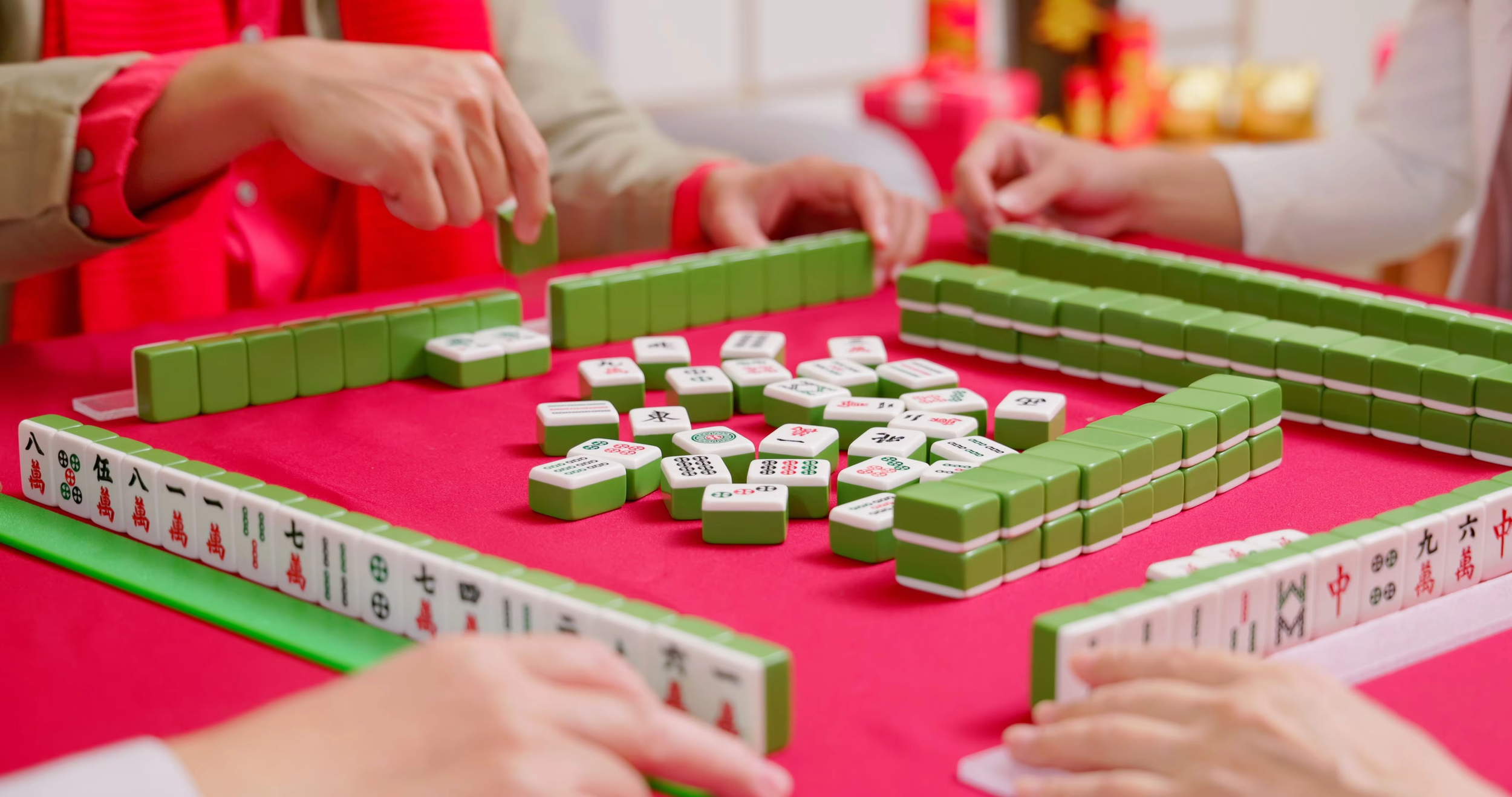 People playing a game of Mahjong at a red table with green tiles and white and patterned tiles.