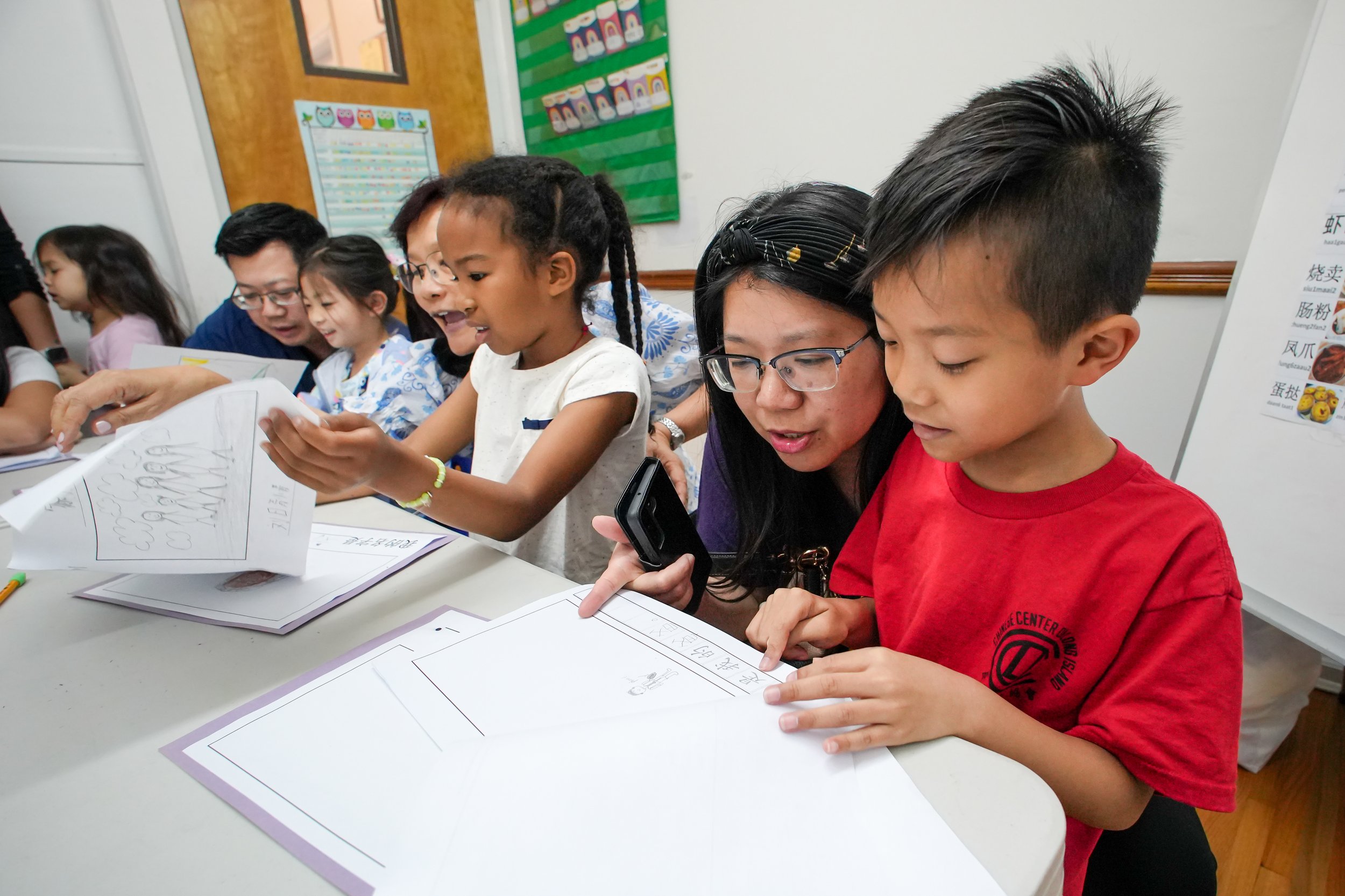 A group of children and adults gathered around a table, looking at papers and sharing a moment of learning in a classroom.