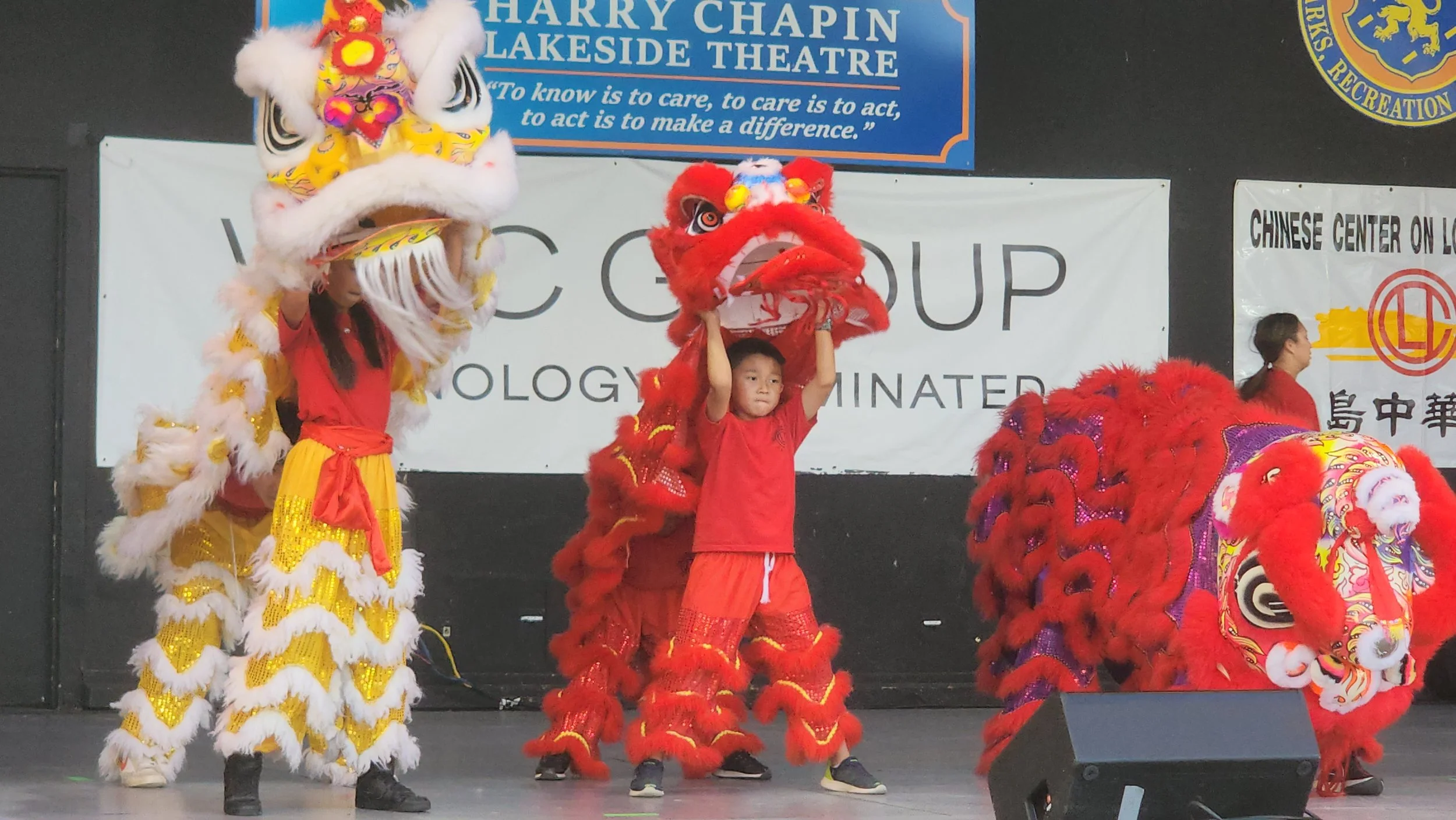 Children performing a lion dance on stage during a cultural event, with colorful lion costumes and background banners.
