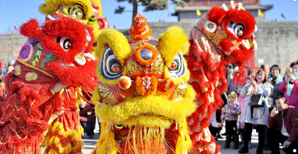Colorful lion dance costumes in red, yellow, and orange with intricate designs, performing in front of a crowd outdoors during daytime.