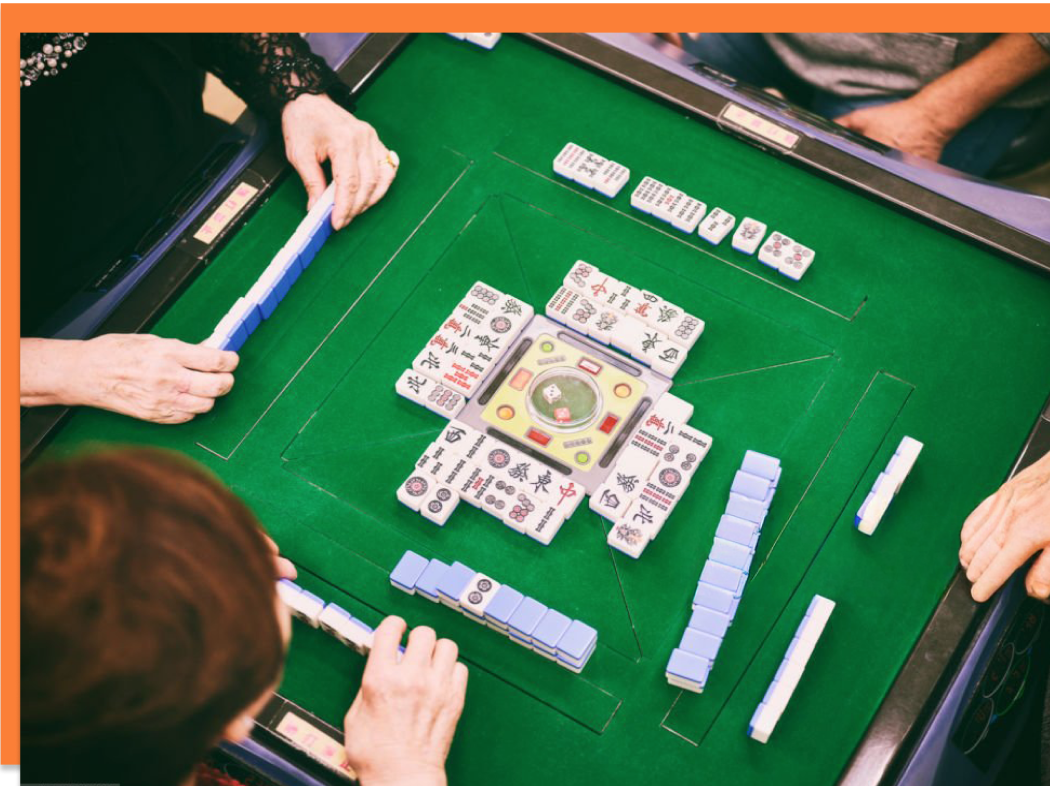 A group of people playing Mahjong around a green felt table, with Mahjong tiles and a spinner in the center.