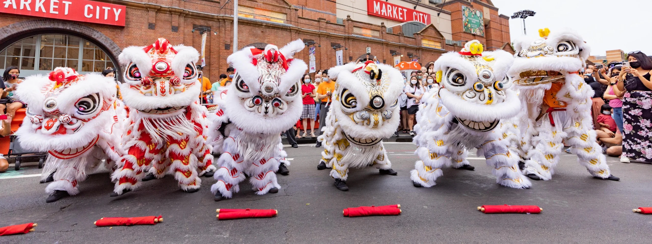 Lion dancers performing at a street parade in front of a crowd outside a brick building with red signage reading 'Market City'.