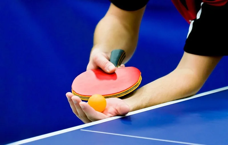 A person playing table tennis, holding a paddle in one hand and a ping pong ball in the other, by the blue table.