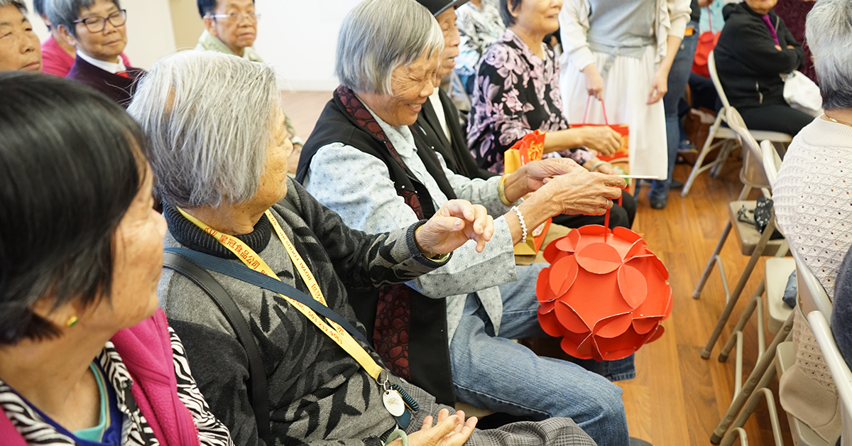 A group of elderly Asian women sitting in chairs, attending a social gathering, with one woman holding a large red decorative paper lantern.