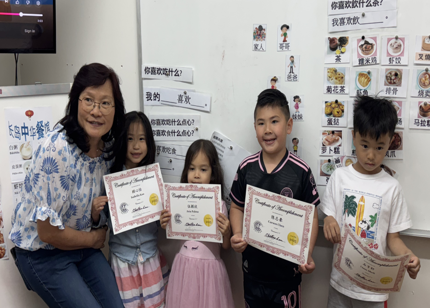 A woman and four children are standing together in a classroom, holding certificates of achievement. The whiteboard behind them has Chinese characters and pictures of food and children.