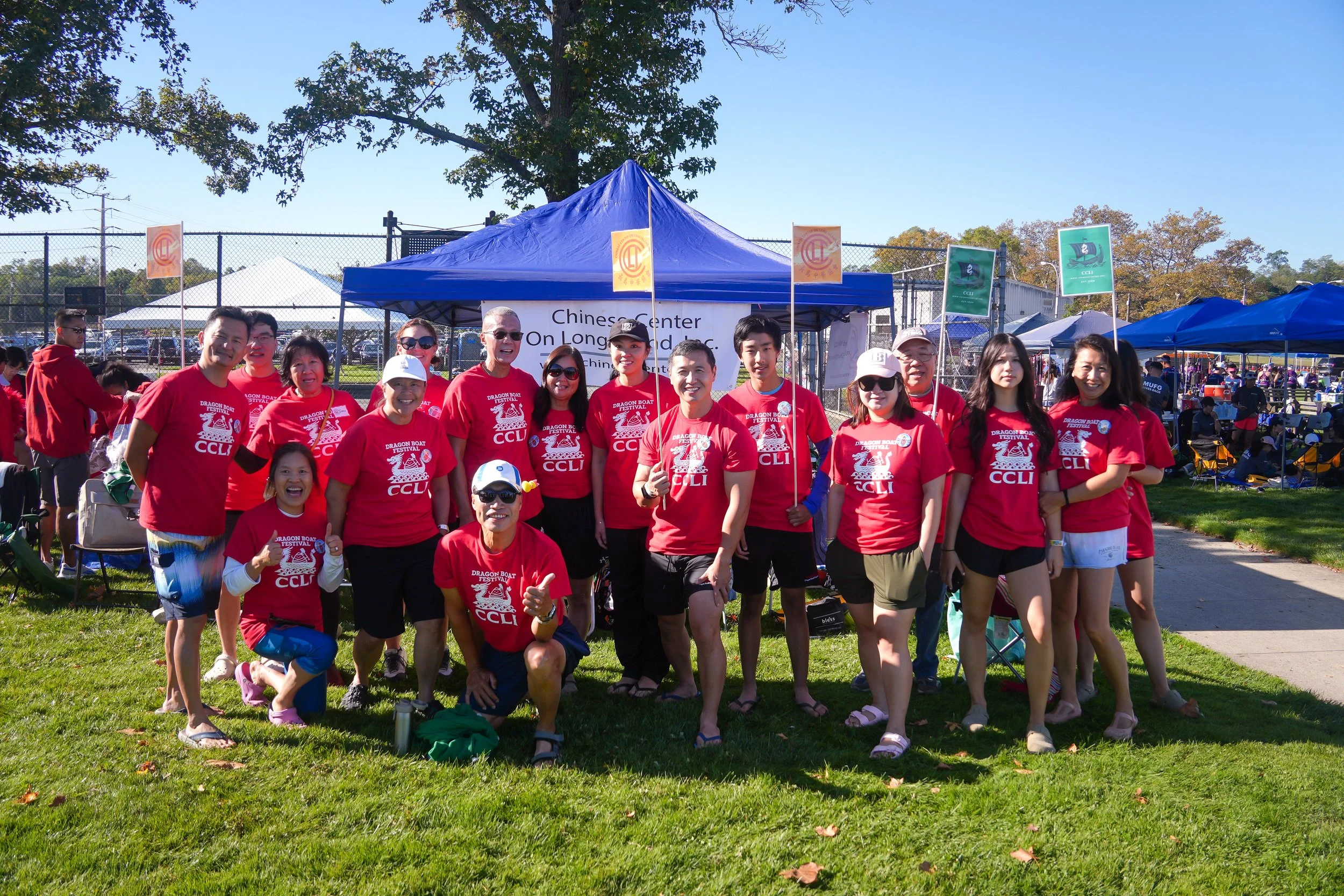 Group of people wearing red shirts at a festival or event outdoors on a grassy area, with tents and banners in the background.