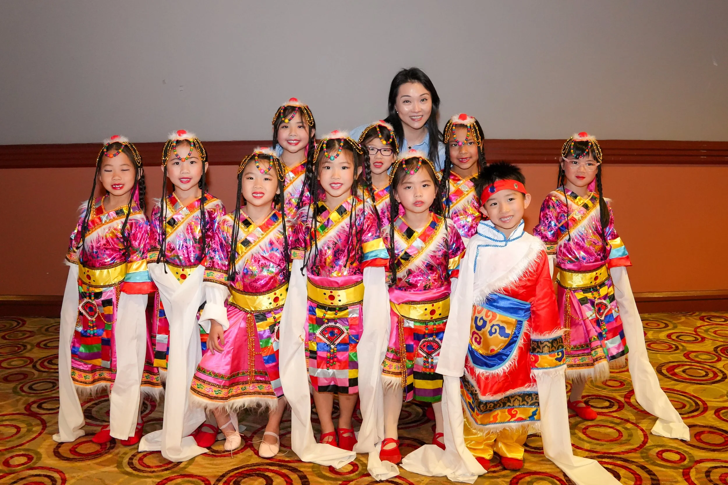 A group of children in traditional Chinese costumes gathered with a woman, posing for a photo indoors on a patterned carpet.