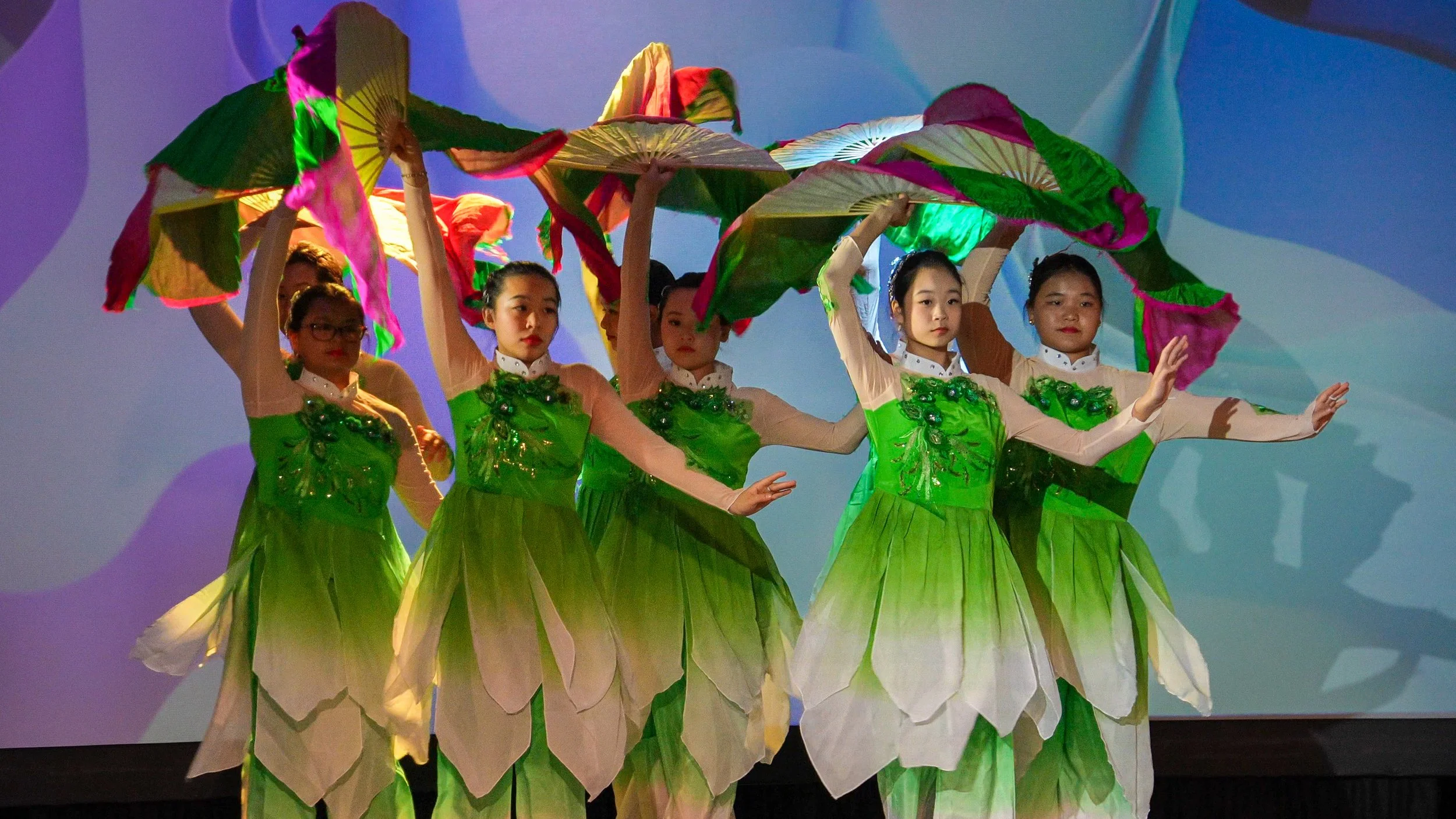 Group of young girls performing a dance on stage, dressed in green costumes with petal-like skirts and holding large, multicolored umbrellas.