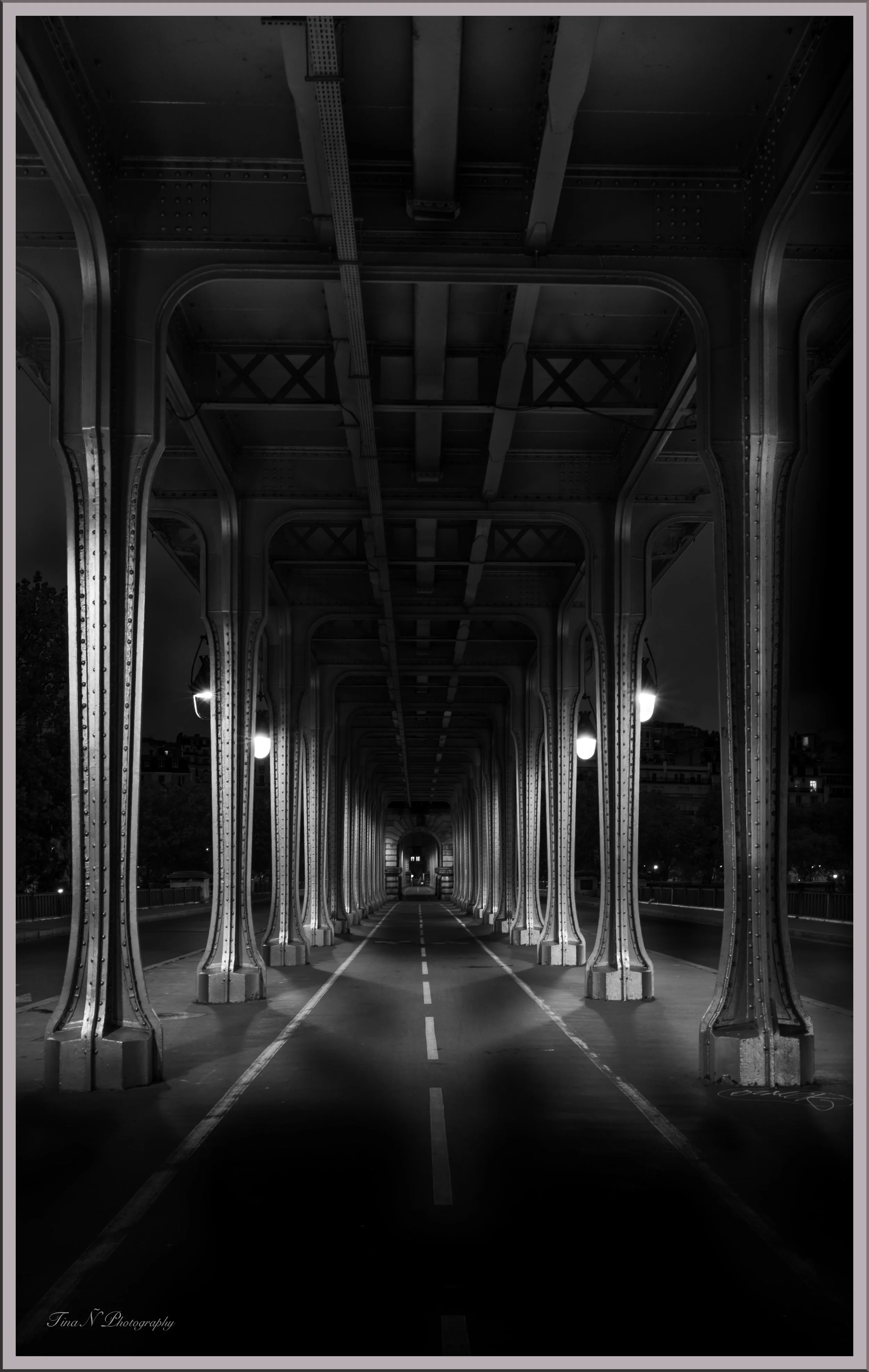 Nighttime view of a metal bridge with a tunnel-like structure, illuminated by several lamps, in black and white.