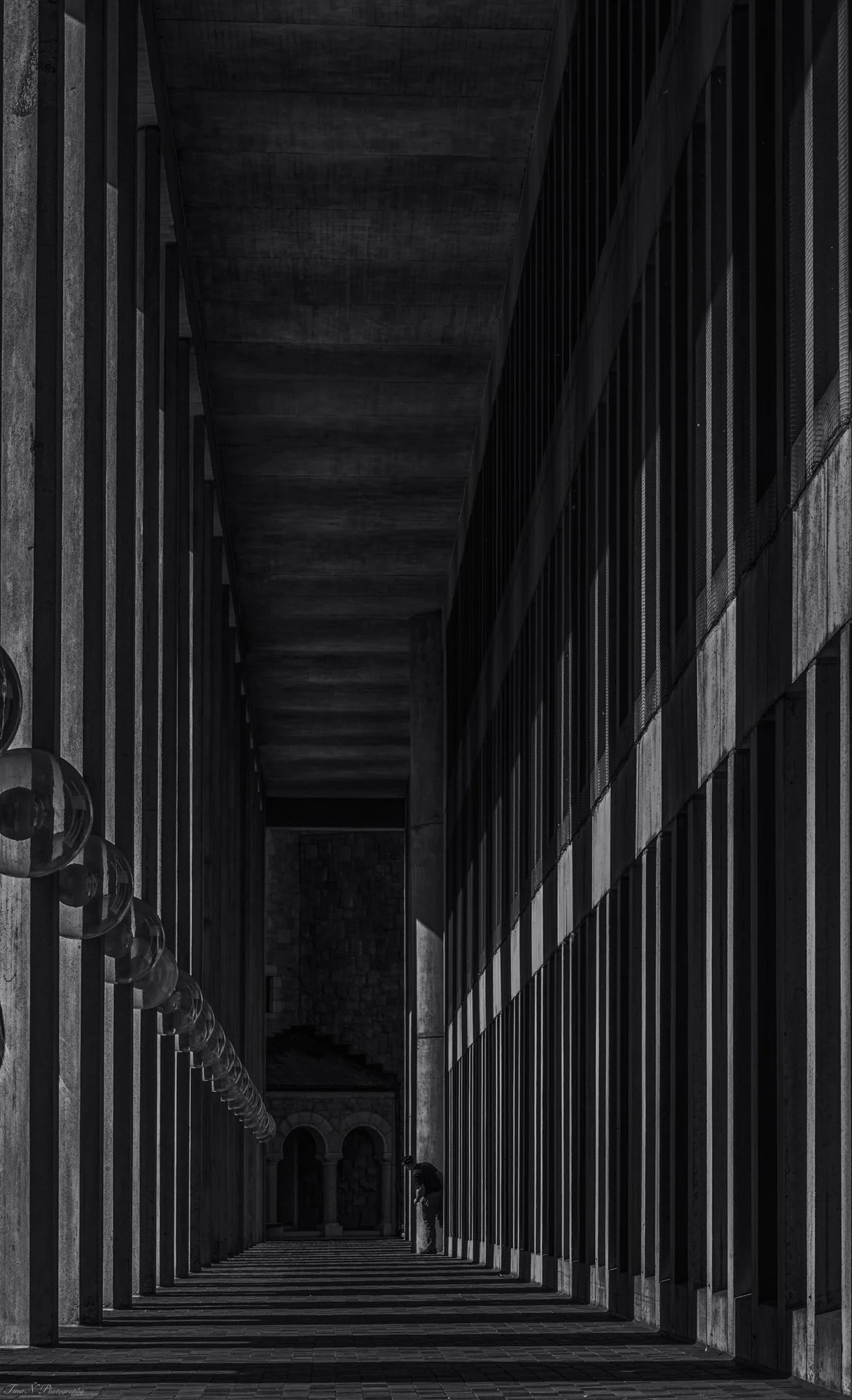 A black and white photo of a modern architectural corridor with tall vertical beams casting striped shadows on the ground, and a person leaning against a column in the distance.
