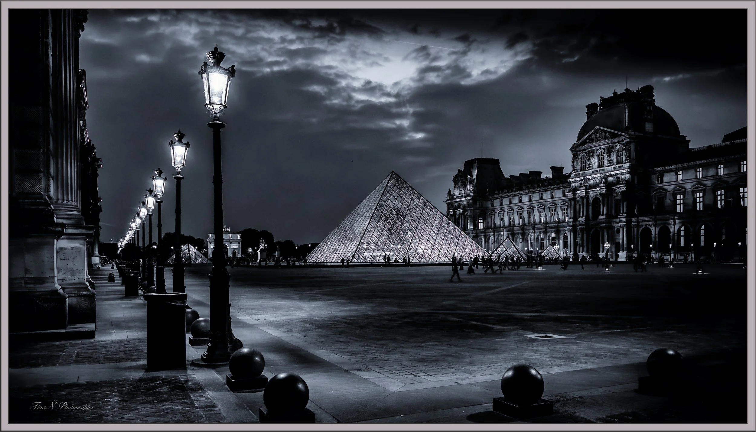 Black and white night photo of the Louvre Museum in Paris with lit street lamps and the glass pyramid entrance, cloudy sky overhead.