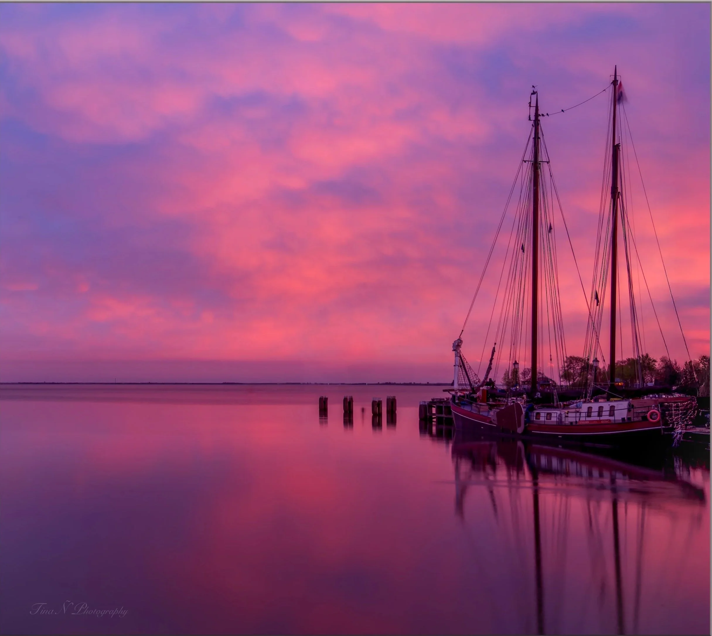 Hoorn, Netherlands. A sunset over calm water with clouds in pink and purple hues. Two sailboats are docked at a pier on the right side, with reflections visible in the water.