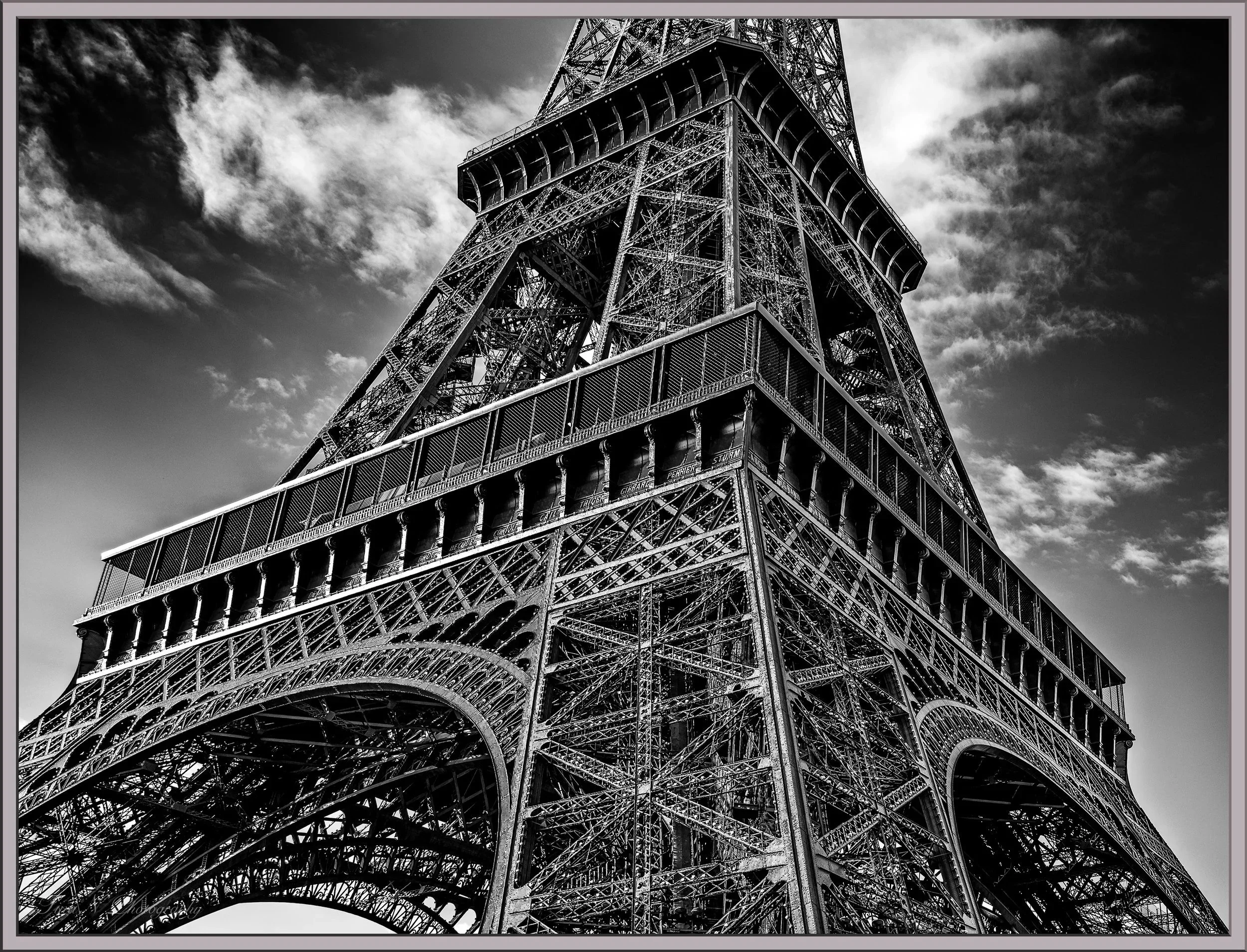 Black and white photograph of the Eiffel Tower viewed from below, emphasizing its intricate iron lattice structure against a cloudy sky.