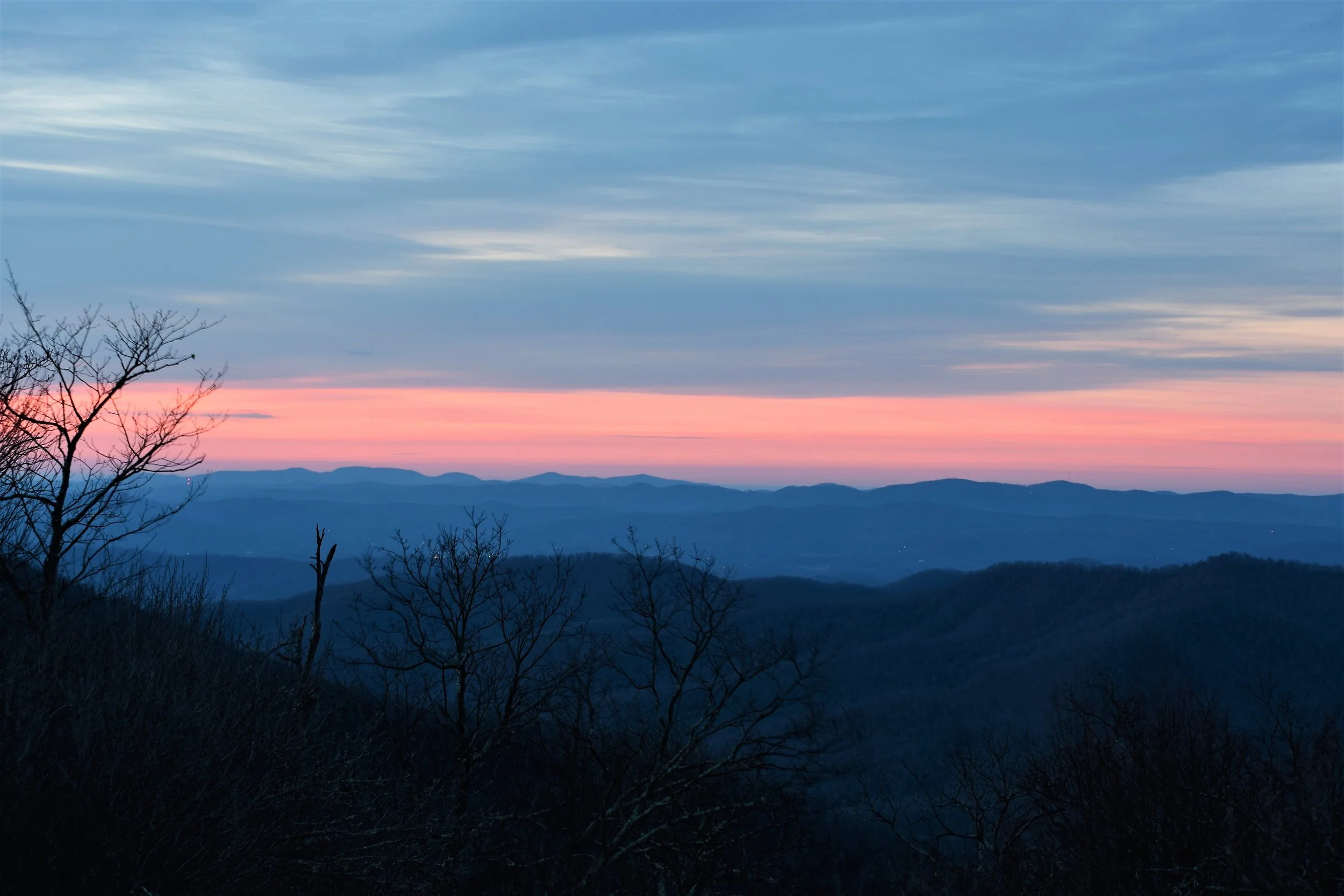 A scenic view of a landscape at dusk with a pink and purple sky, distant mountains, and silhouetted trees in the foreground.