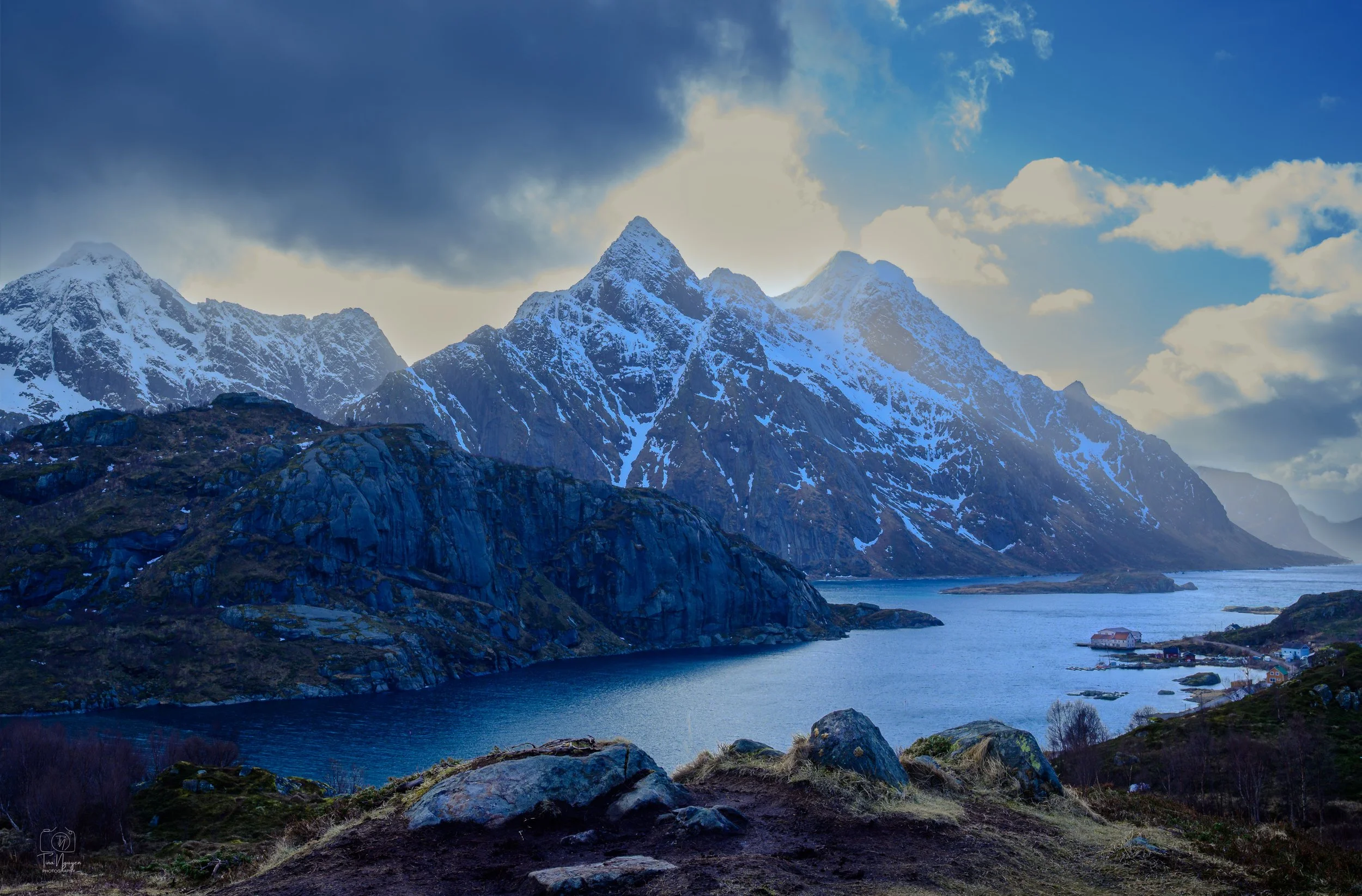 Snow-capped mountains rising above a fjord with a small village at the water's edge and a partly cloudy sky overhead.