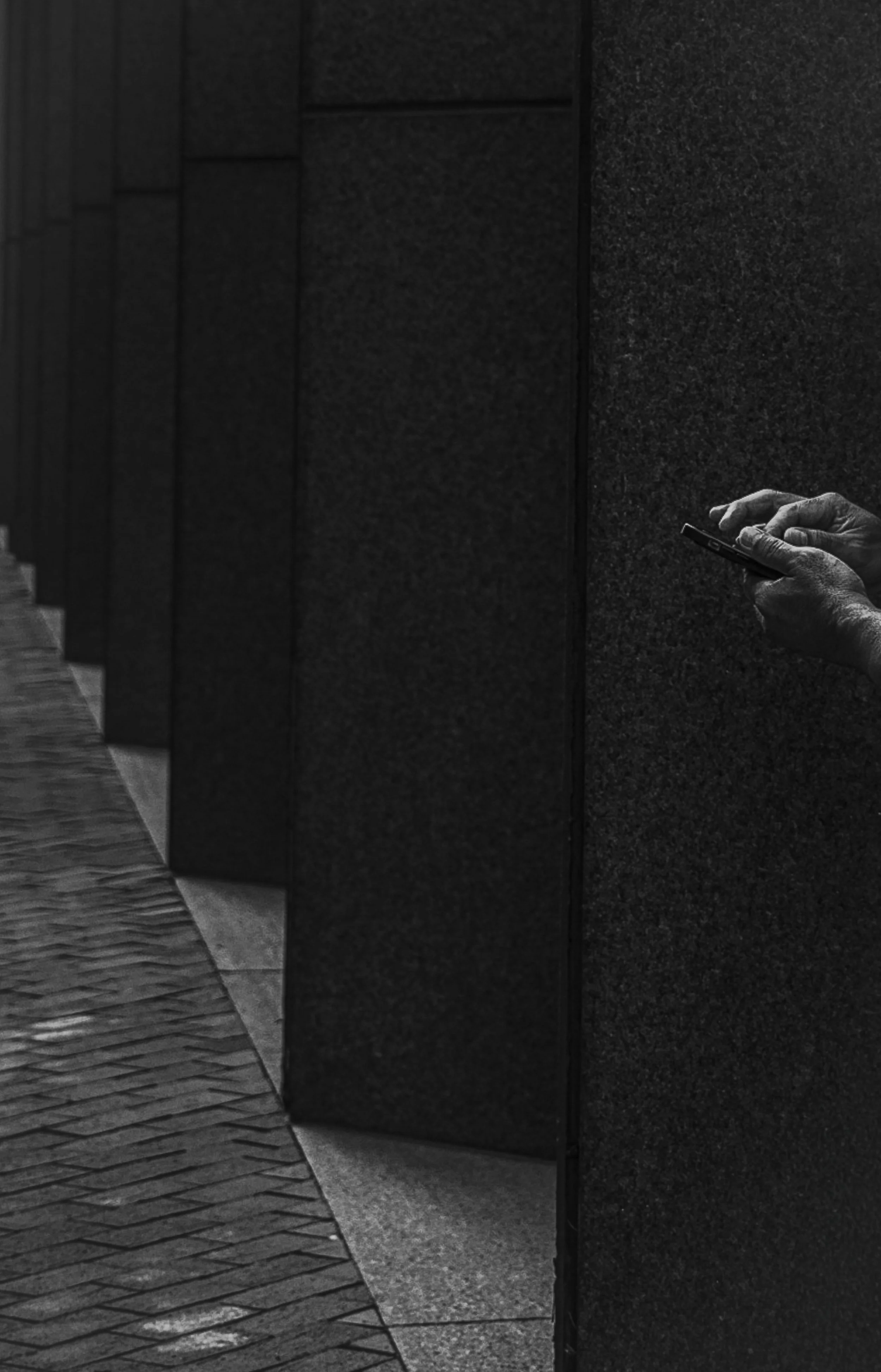 A black and white photo of a hand holding a mobile phone against a dark wall, with a textured sidewalk on the left.