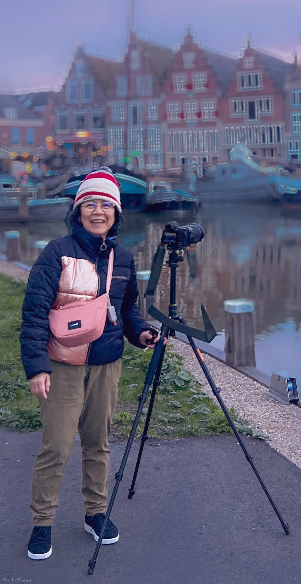 Woman standing next to a camera on a tripod near a waterway with boats and colorful buildings in the background.